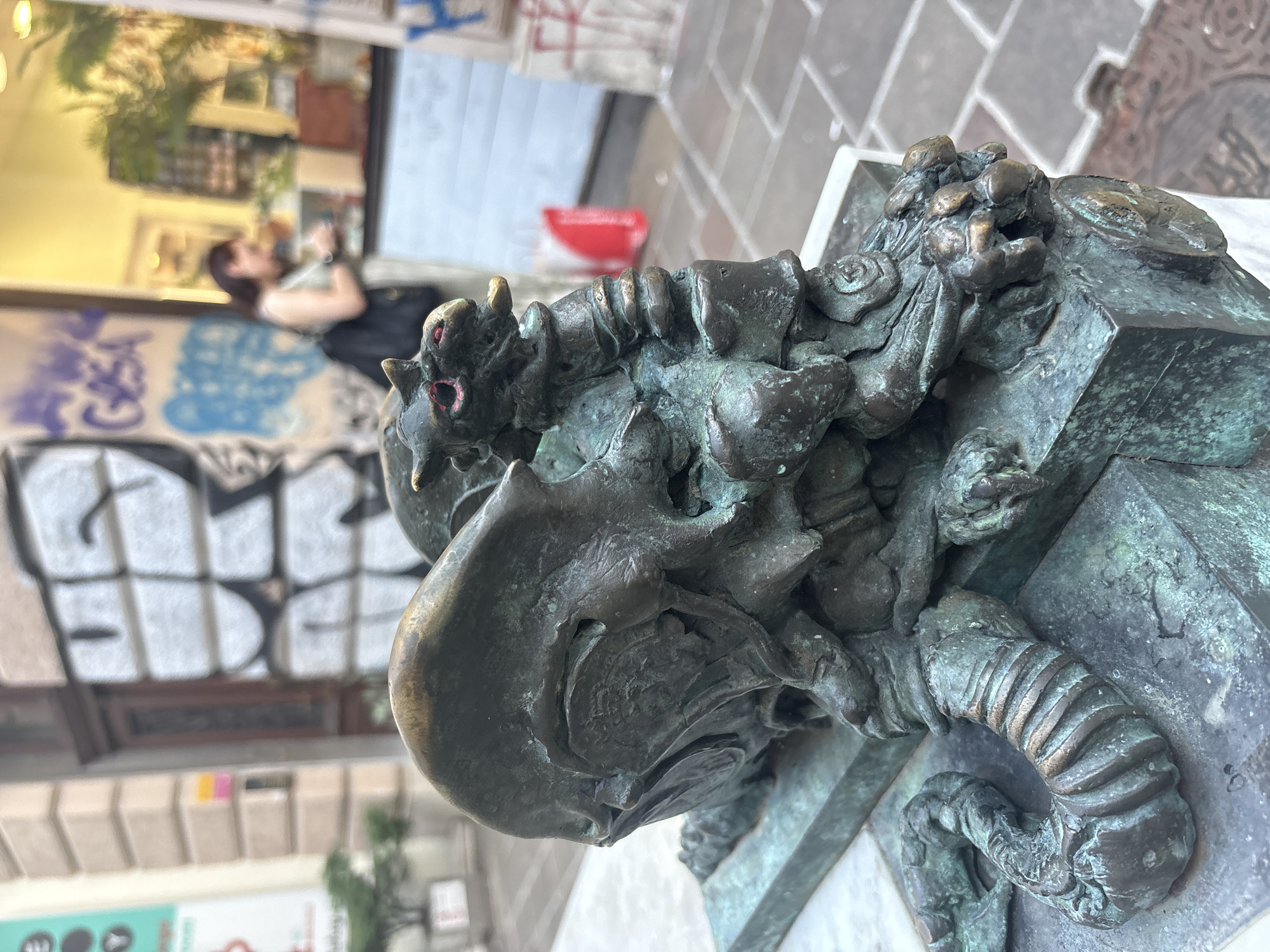 A weathered bronze statue of a dragon, showing signs of patina and age, is positioned in the foreground of the image. The dragon is depicted in a crouched or resting position, with its head turned to the right. Its eyes are painted red, and its mouth is slightly open, revealing a golden tongue. The statue is mounted on a stone base. In the background, a woman wearing a light-colored top and a dark backpack is visible, standing in front of a shop window. The shop window displays various items and is illuminated from within. The street outside is paved with cobblestones. Graffiti is present on a building wall behind the statue and to the left of the woman. The overall scene suggests an outdoor urban environment, possibly a street or a public square.