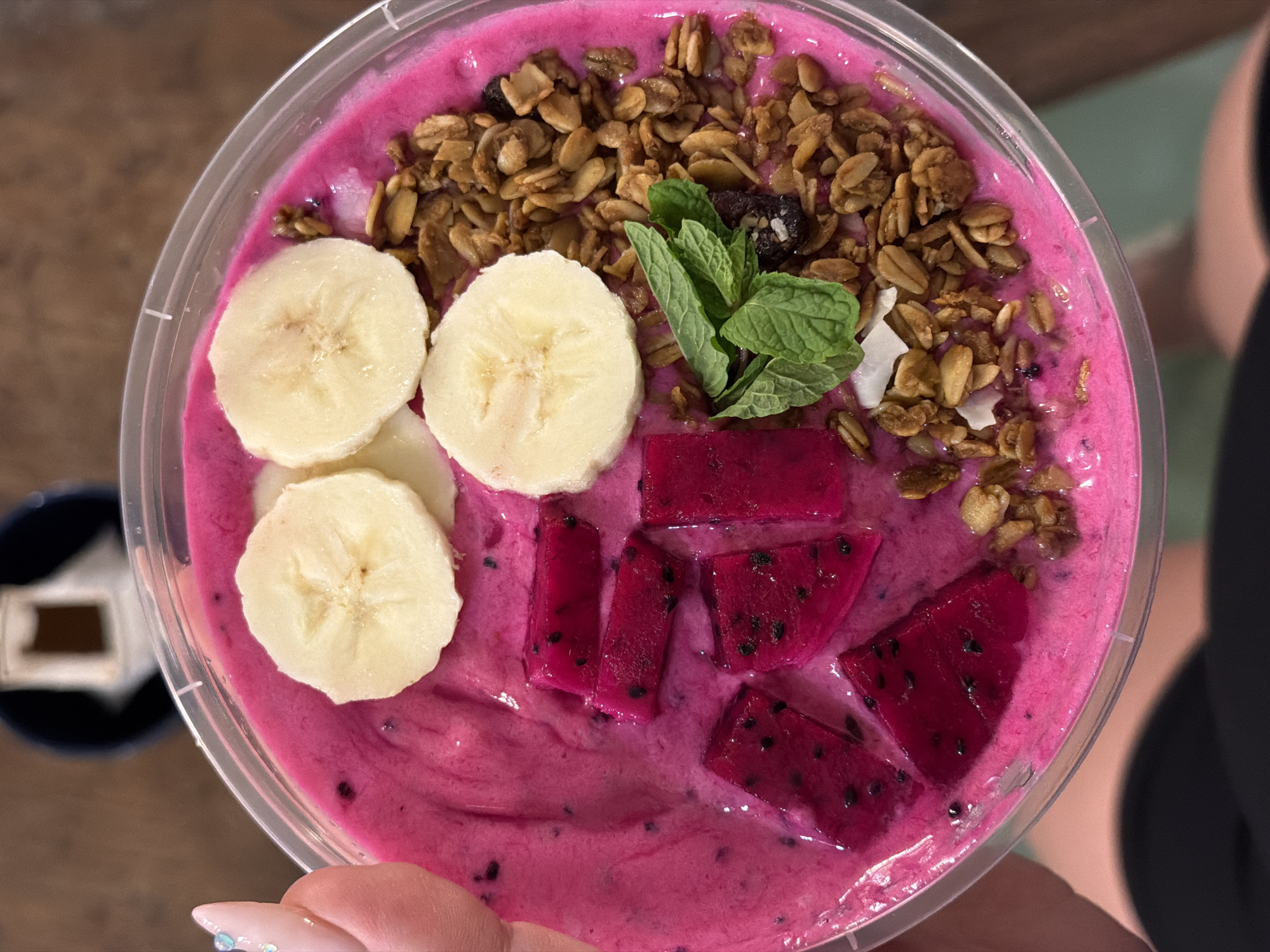 An overhead, slightly angled shot depicts a dragon fruit smoothie bowl presented in a clear, round plastic container, held by a human hand. The smoothie base is a vibrant fuchsia-pink, displaying a smooth, semi-viscous consistency with dispersed small black seeds.
Toppings are arranged across the surface: three circular slices of banana are positioned in the upper-center and mid-right. To the left of the center, several irregular pieces of diced dragon fruit, characterized by their intense dark pink hue and visible black seeds, are partially submerged in the smoothie. The right side of the bowl is covered with granola, appearing as clusters of golden-brown oats and grains. Within the granola, sporadic white flakes, resembling coconut, and a dark, shriveled piece of dried fruit are discernible. A small sprig of fresh green mint leaves is placed atop the granola in the mid-right section.
A human hand, featuring light pink or nude nail polish with minor glitter or rhinestone accents on the thumb, grips the clear container from the left side. A portion of the individual's arm and shoulder, clad in dark fabric, is visible below the container.
The background is intentionally blurred, revealing a warm brown surface, consistent with wood, in the upper portion of the image. A dark blue or black object, possibly a cup or bowl, containing a white insert with a small brown, rectangular element on top, is situated in the upper background. A light green or teal surface is visible in the bottom right corner of the frame. The scene is located in North Kuta, Indonesia.
