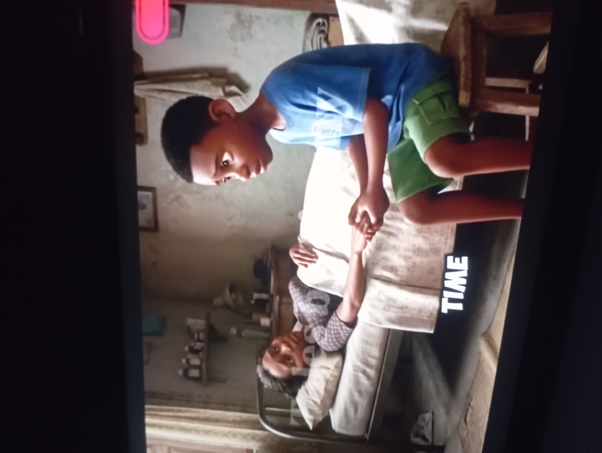 A boy in a blue shirt and green shorts sits on a chair, leaning forward with his hands on a person lying on a bed. The person on the bed is covered with a white sheet and has their head resting on a pillow. "TIME" is written on the side of the bed. The room is cluttered with shelves and a fan. The scene is located in the city of Unknown, Unknown.