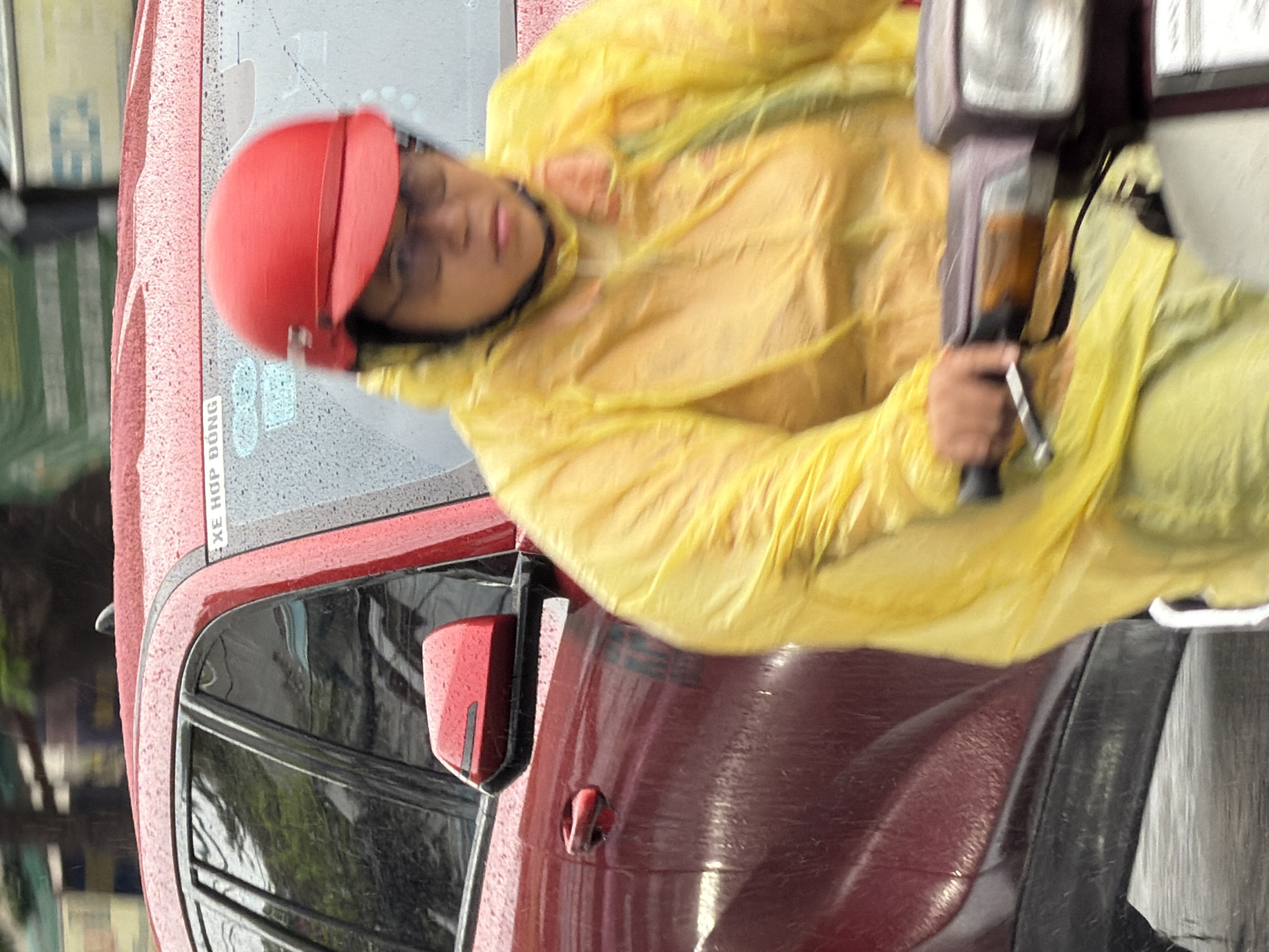 In Nha Trang, Vietnam, a person wearing a bright yellow raincoat and a red helmet is riding a scooter on a wet road. Raindrops are visible on the person's clothing and on a red car parked nearby. The car has tinted windows and a side mirror. The overall scene suggests a rainy day in an urban environment.