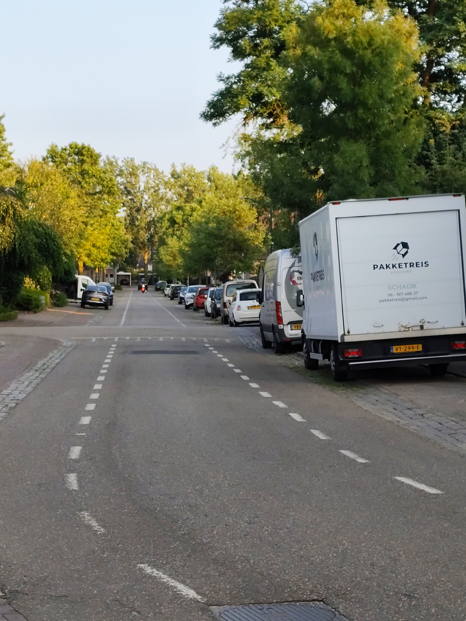 A white delivery truck with the logo "PAKKETREIS LOGISTIEK" is parked on the right side of a paved road lined with trees. The truck has a license plate that reads "VT-299-F". Next to it, a white van is also parked. Several other cars and a scooter are visible further down the road. The scene appears to be in a residential area in Schaijk, Netherlands, characterized by lush green trees and a clear sky. The road has white dashed lines indicating a lane divider.