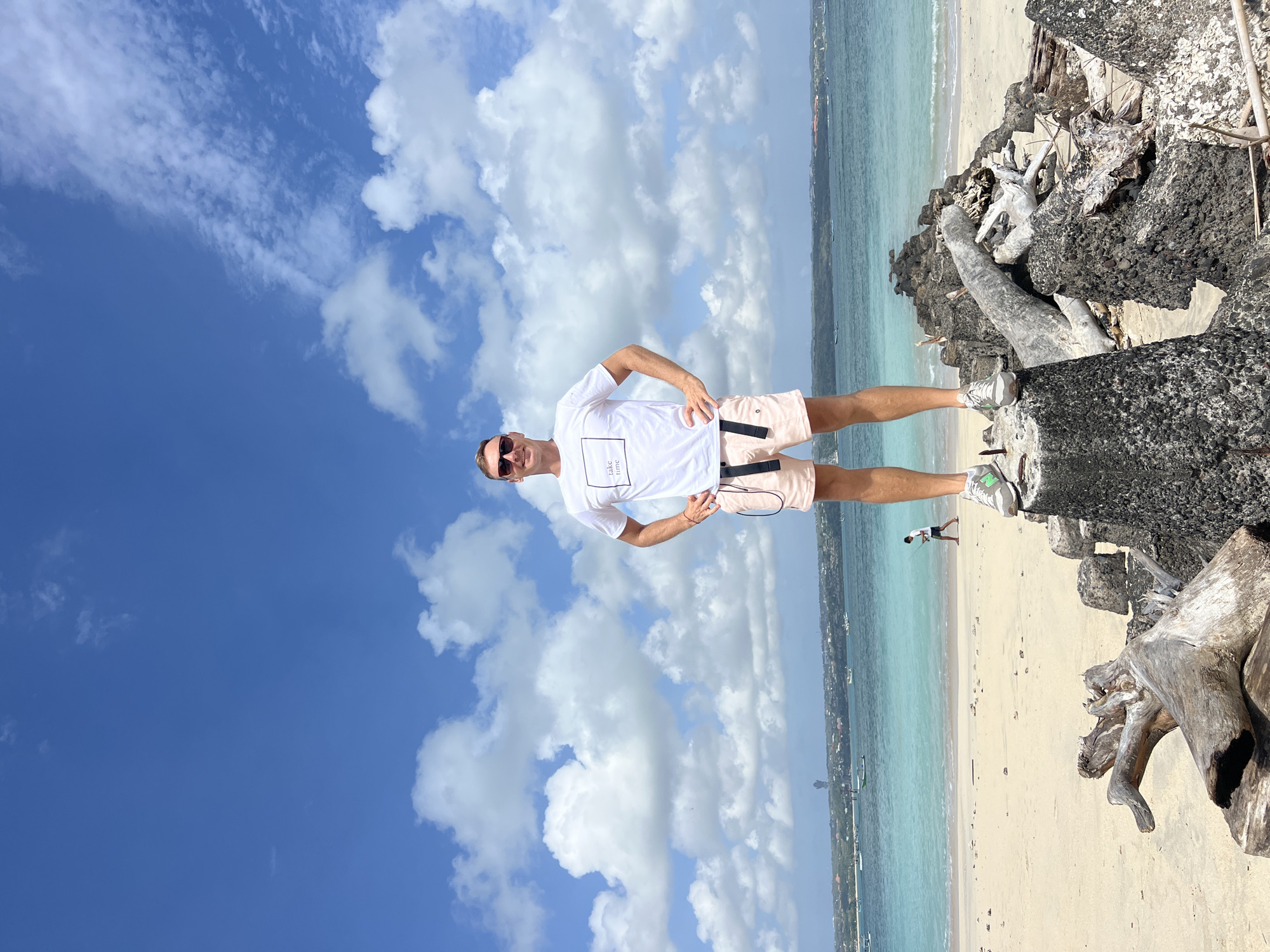 A man stands on a pile of rocks and driftwood on a beach in Denpasar, Indonesia, with the ocean and sky in the background. The man is wearing a white t-shirt with text that reads "take time", pink shorts, sunglasses, and grey sneakers. He is looking at the camera with his hands on his hips. The beach is sandy and the water is turquoise. There are boats in the distance and a person walking on the beach. The sky is blue with white clouds.