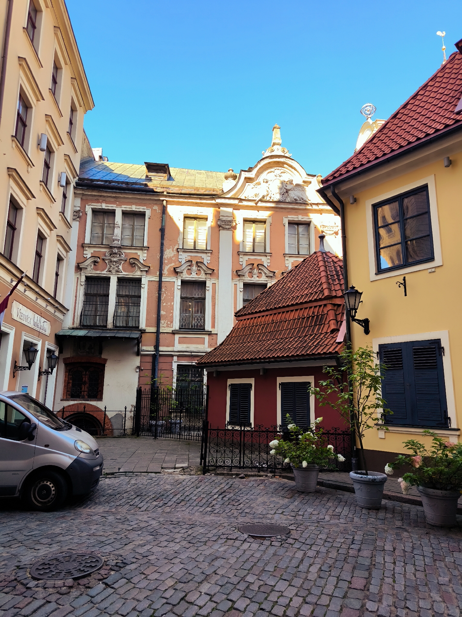 The image shows a narrow, cobblestone street in Riga, Latvia, likely in the Old Town, with historic buildings on either side under a clear blue sky. On the left, a light-colored building with multiple windows and decorative facade elements stands adjacent to a darker building with ornate window frames and a sign that reads "Viesnīca Hotel de Roma." A silver van is parked on the street, facing towards the left edge of the frame. In the center of the image, a small, red building with a red-tiled roof and dark shutters is nestled between taller structures. A black metal fence separates this area from the street. On the right, a yellow building with a dark-tiled roof features a large window reflecting the surrounding architecture and a dark shuttered window on the lower level. Potted plants with white flowers are placed in front of the red and yellow buildings. The cobblestone pavement is well-maintained, and the overall scene depicts a charming, historic urban environment.
