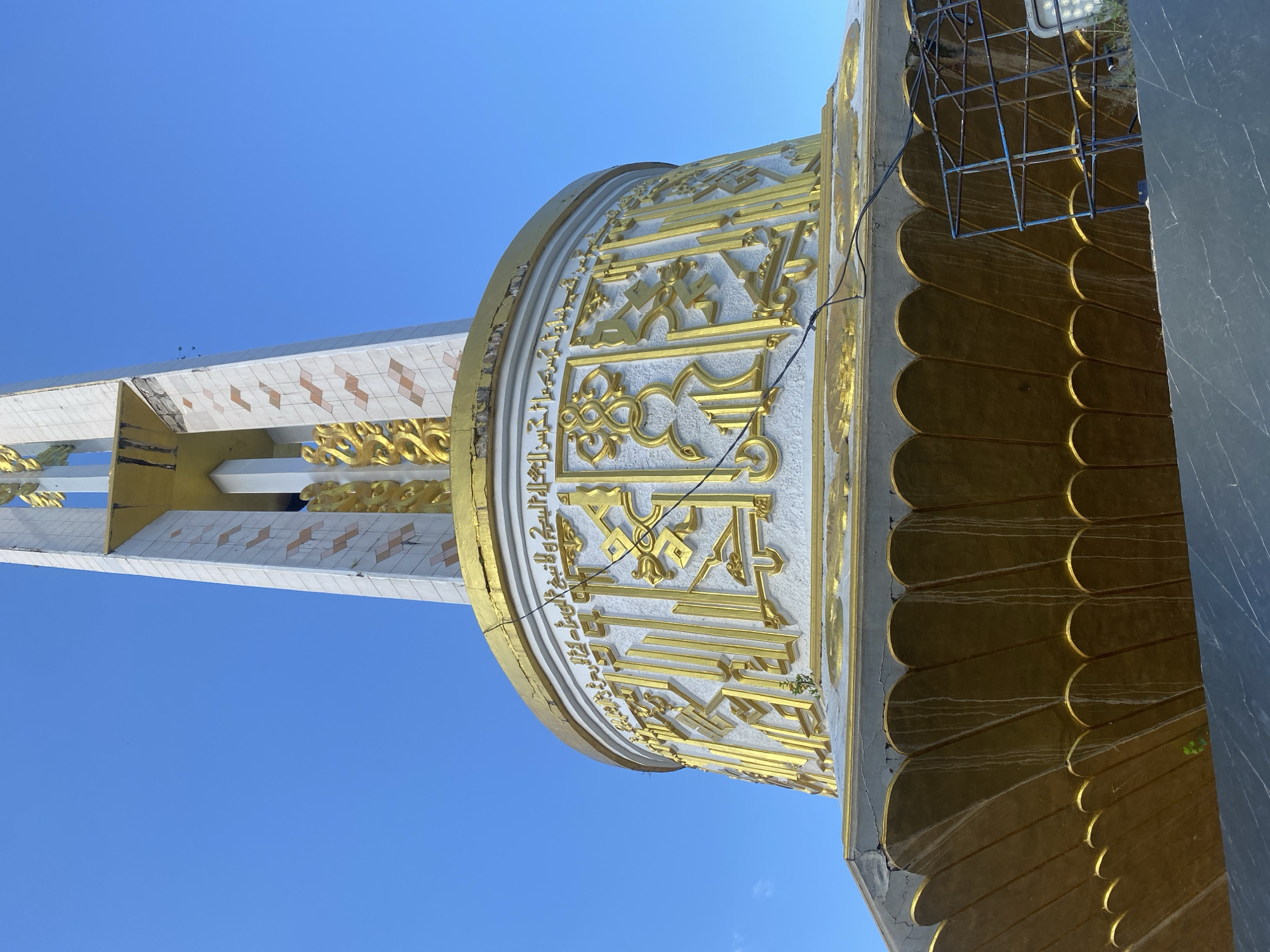 The image shows a low-angle view of the upper portion of a monument, likely a minaret or obelisk, against a clear blue sky. The monument is predominantly white with decorative gold elements and Arabic calligraphy.
The base of the monument is a wide, octagonal structure with a decorative band of gold, shaped like overlapping petals or leaves. This band is stained with vertical streaks, suggesting water or weathering. Above this is a cylindrical section, also white, adorned with intricate, raised gold calligraphy. The calligraphy is densely packed and appears to be Arabic script, though the specific text is illegible in detail. The gold work is ornate and stylized.
Continuing upwards, the monument narrows into a tall, rectangular shaft. This shaft is clad in white tiles, some of which are arranged in a diamond pattern with lighter brown or reddish accents. There are visible signs of wear and tear on the tiling, with some sections chipped or missing, revealing underlying material. Vertical gold decorative elements are also integrated into the sides of this shaft. A portion of what appears to be scaffolding or a temporary support structure, made of blue metal bars, is visible on the right side of the frame, obscuring part of the monument's base.
The overall impression is of a significant religious or civic structure, possibly undergoing maintenance or showing signs of age. The clear sky suggests it is daytime, and the upward angle implies the monument is quite tall.
This monument is located in Martapura, Indonesia. Specifically, it is the Tugu Martapura, also known as Monumen Perjuangan (Monument of Struggle), located in Martapura, Banjar Regency, South Kalimantan, Indonesia.
