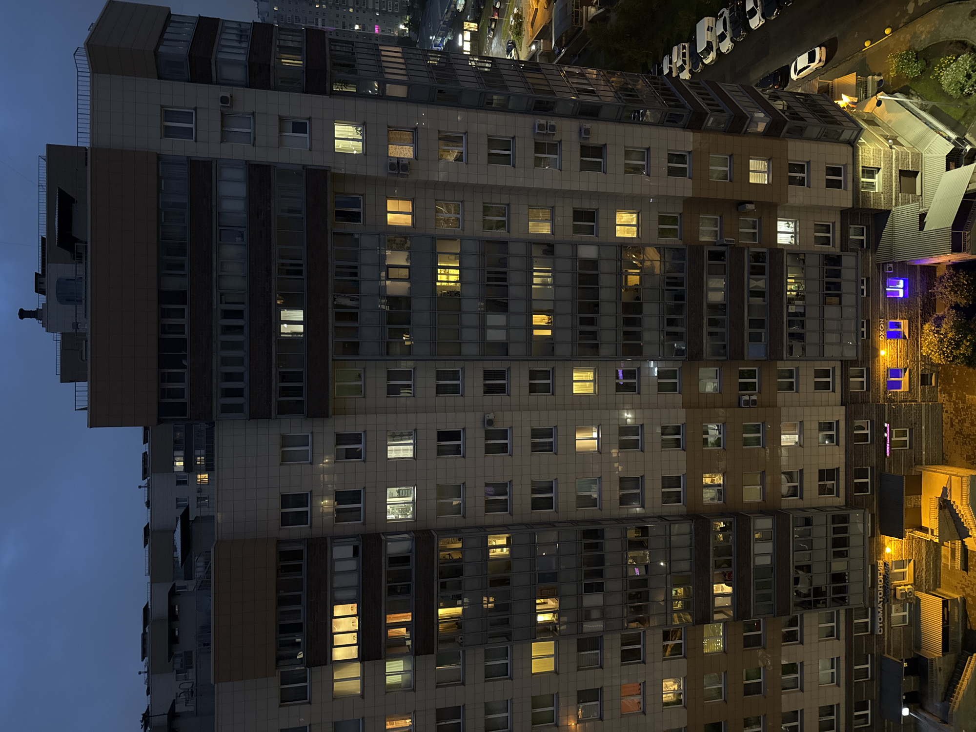 The image depicts an exterior view of a tall, multi-story apartment building in an urban setting at twilight. The building is primarily clad in beige and brown panels, with numerous windows, many of which are illuminated from within, suggesting occupied apartments. Some windows are dark. The architecture features protruding balconies and a distinct angular design.
At the base of the building, there are entrances with awnings and visible signage, including "СТОМАТОЛОГИЯ" (Dentistry) and "OZON," which is likely a retail outlet. A street runs alongside the building, with several parked cars, including a white van, white sedan, and other vehicles. Streetlights and the lights from the building cast an ambient glow on the surroundings.
The sky above is a deep twilight blue, indicating the time is either early evening or late dusk. The overall scene suggests a residential neighborhood in a Russian city, possibly in the midst of a transition from day to night. No people are clearly discernible within the apartments or on the street.