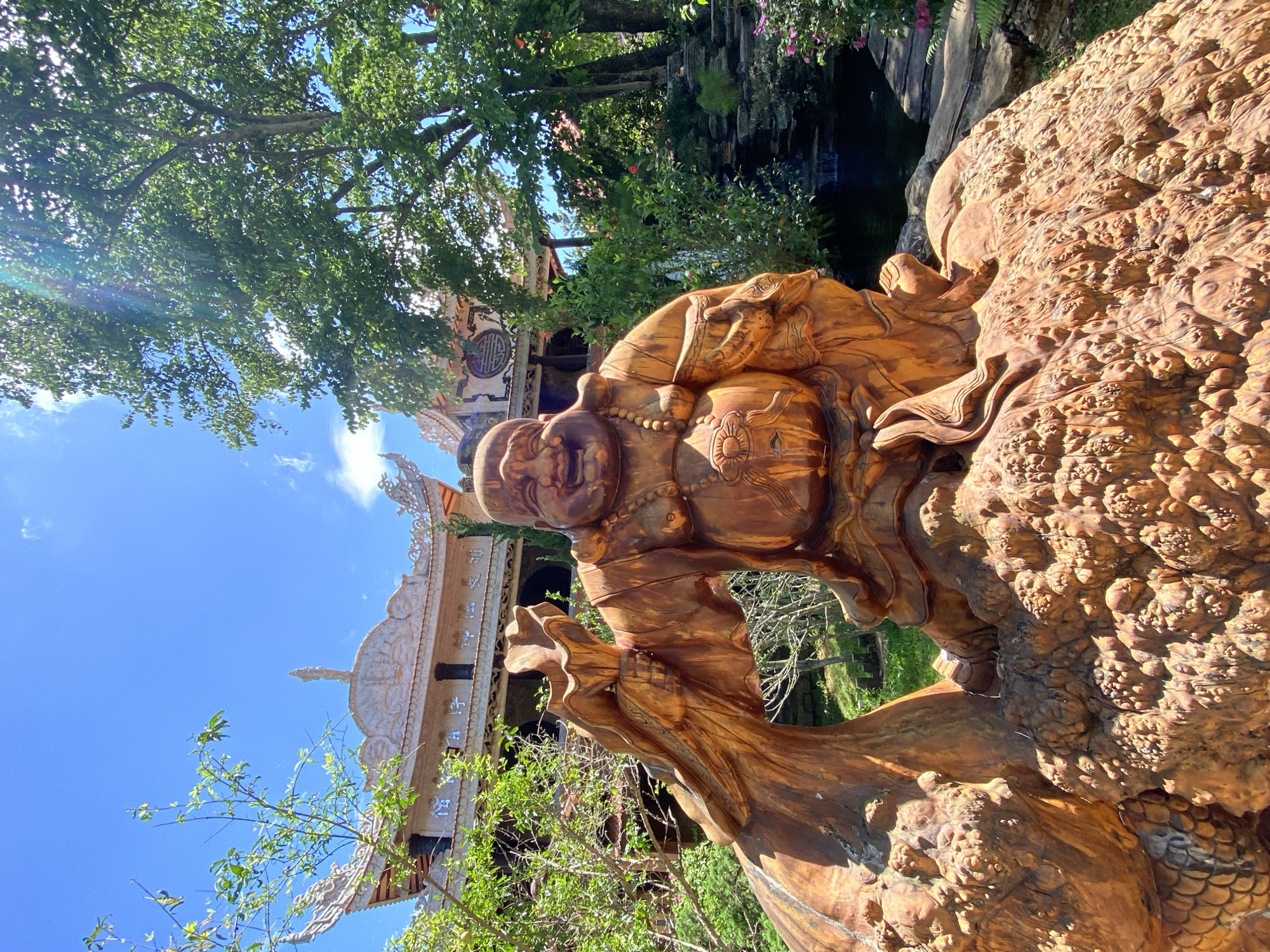 This media file captures a close-up view of a large, intricately carved wooden sculpture of a jovial, perhaps Buddhist, figure. The sculpture is rendered in warm, reddish-brown tones, with detailed textures suggesting flowing robes and beaded necklaces. The figure's face is round and smiling, with closed eyes, conveying a sense of peace and contentment. The sculpture is positioned outdoors, with lush green foliage and trees forming a backdrop.
Behind the sculpture, a portion of a building with ornate, traditional architecture is visible. The building features multiple tiered roofs with decorative trim and what appear to be carved motifs. The overall scene is bathed in bright sunlight, suggesting it is daytime. The sky is a clear, vibrant blue with scattered white clouds, indicating pleasant weather.
The lower right of the image focuses on a textured section of the sculpture that resembles scales or a rough, organic surface, possibly part of the larger structure the figure is integrated with. The wooden carving appears to be a significant artistic or religious artifact, possibly situated within a temple, garden, or other culturally significant site. The image's orientation is vertical, suggesting it was taken with the camera held in portrait mode. The scene evokes a sense of serenity and appreciation for craftsmanship. There is no visible text in the image. The location context provided is Thôn 5, Vietnam, which aligns with the architectural style and potential cultural subject matter.