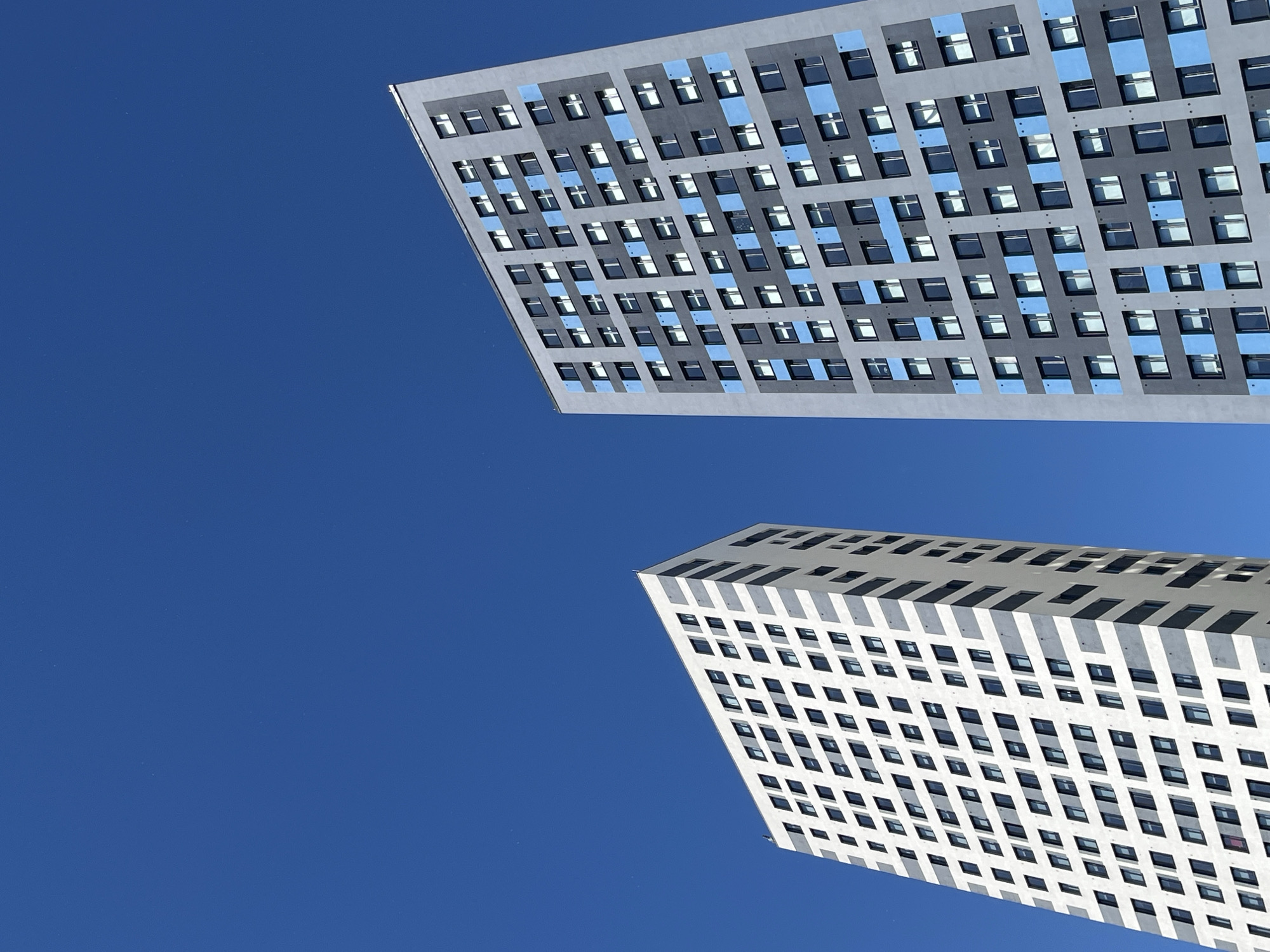 This media file is a low-angle shot of two modern buildings against a clear blue sky. The building in the foreground is a white, multi-story structure with a repeating pattern of dark square windows and diagonal white accents. The building in the background is a taller, darker structure with a grid of evenly spaced windows, some of which are highlighted with blue accents. Both buildings appear to be residential or office complexes, characterized by their repetitive architectural elements. The clear blue sky suggests it is daytime, likely sunny, with no clouds visible. The architecture suggests an urban environment, possibly a city with contemporary development. The perspective of the shot, looking upwards, emphasizes the height and scale of the buildings and creates a sense of looking into the sky. There is no visible text in the media file. The scene is devoid of people or any discernible activity, focusing solely on the architectural forms and the sky. The overall impression is one of modern urban design against a vast, open sky.