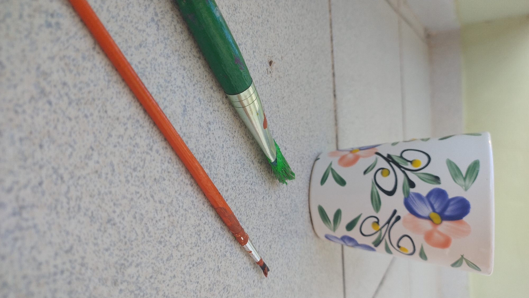 Two paintbrushes and a decorated mug are placed on a light-colored, speckled tiled floor. The mug, positioned in the background and slightly to the right, is white with hand-painted blue and pink flowers and green leaves. One paintbrush, with a green handle, is in the foreground, lying horizontally across the frame. Its bristles are coated with green paint. The second paintbrush, with an orange handle, is positioned below the first, diagonally from the bottom left corner towards the center. Its bristles appear to be tipped with a reddish-brown paint. The scene is lit from the side, casting subtle shadows. There are no people present, and the setting appears to be indoors, possibly in a home or studio. The location is not specified as being in Perea, Greece.