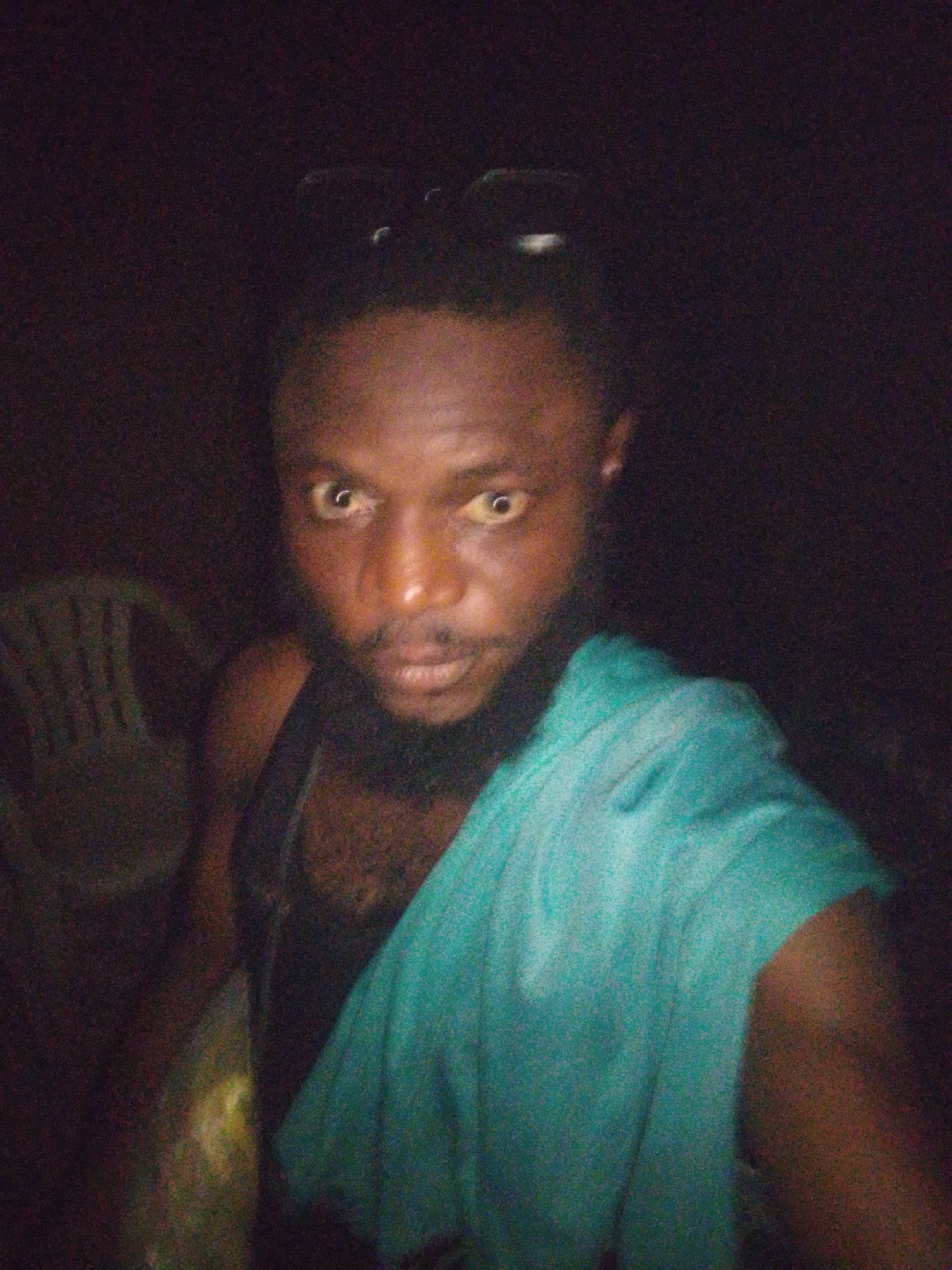 The image depicts a man with dark skin, a beard, and short, dark hair, wearing a black tank top and a turquoise cloth draped over his shoulder. He is looking directly at the camera with a serious expression. The background is dark and indistinct, with a white plastic chair partially visible to the left. The location is reportedly Lokoja, Nigeria, but the lack of environmental details makes verification impossible from the image alone.