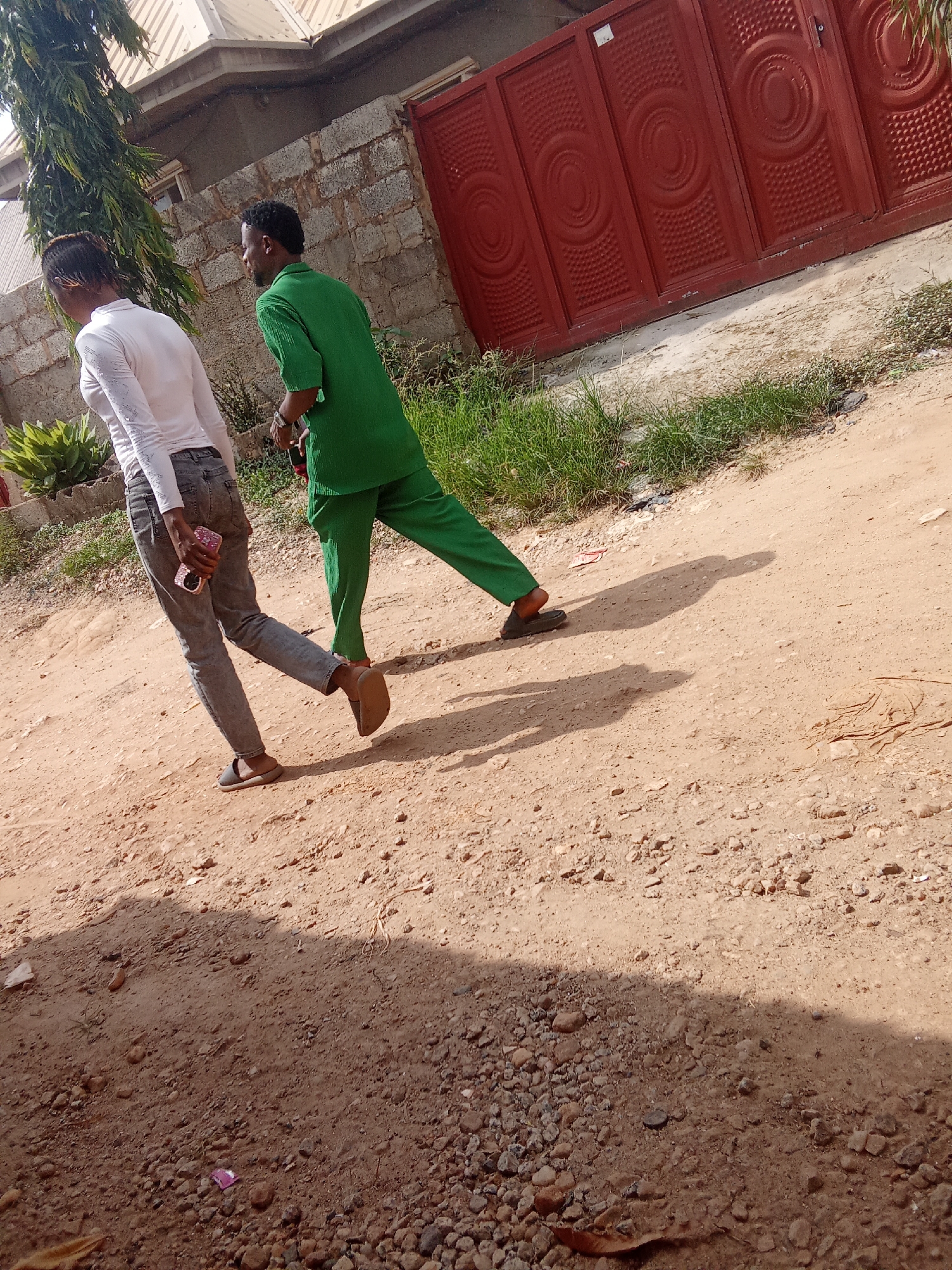 The image shows two individuals walking on a dirt path in Kagini, Nigeria. The person in the foreground, on the left, is wearing a white long-sleeved shirt and gray jeans, and is holding a pink phone. They are walking away from the camera, with their head turned slightly to the right. The second person, on the right, is wearing a green shirt and green pants, and is also walking away from the camera. They are positioned behind and slightly to the right of the first person.
The setting appears to be outdoors, with a building constructed of concrete blocks and a large red metal gate visible in the background. Green vegetation, including trees and grass, is also present. The ground is covered in dirt and small stones. The lighting suggests it is daytime, likely sunny, as evidenced by the strong shadows cast by the individuals. There is no visible text in the image. The overall impression is of a casual daytime stroll in a residential or semi-urban area in Nigeria.