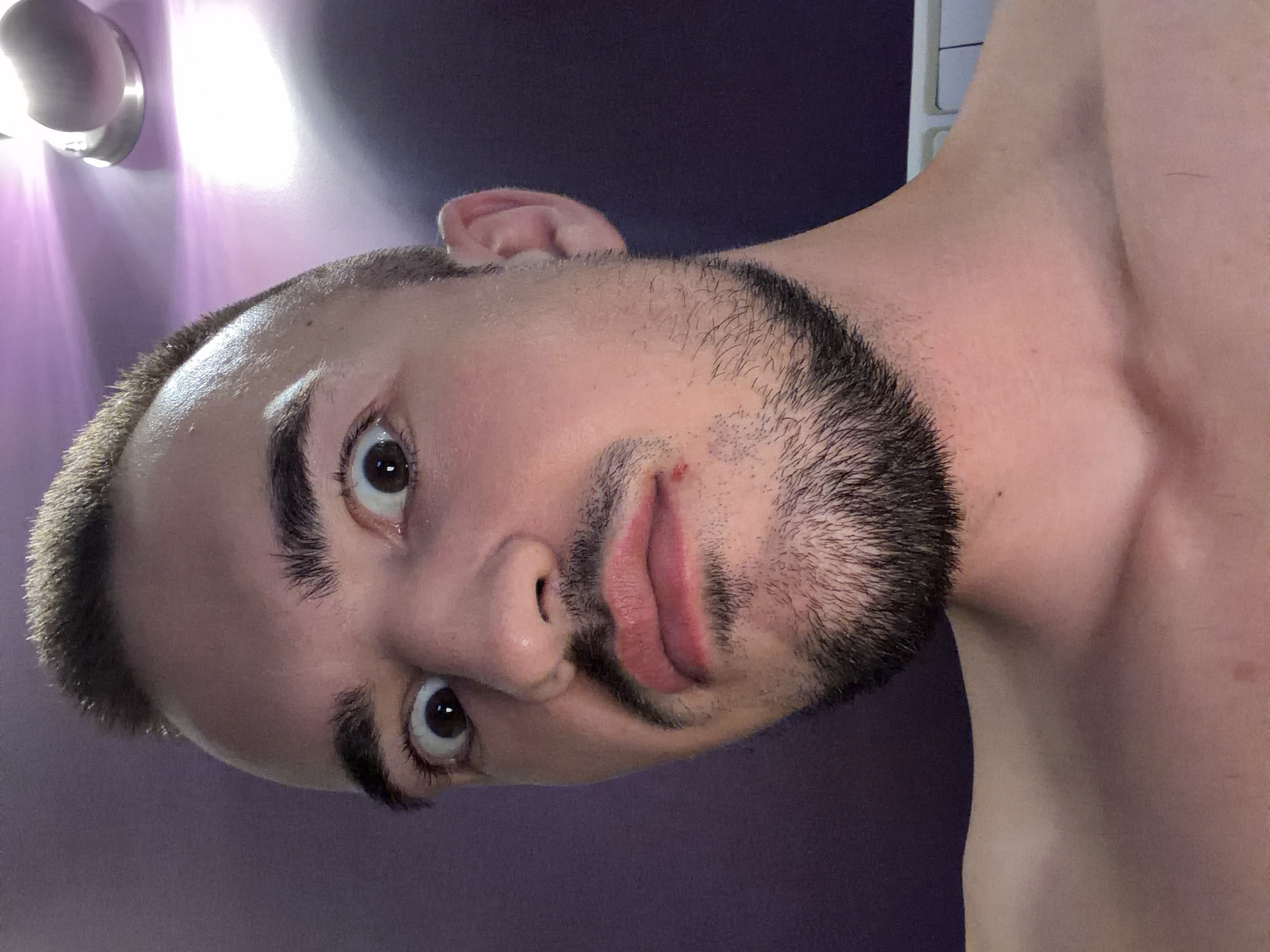 The image shows a close-up, forward-facing selfie of a man with a surprised or wide-eyed expression. He has short, spiked brown hair, dark eyebrows, and a goatee and mustache. His lips are slightly parted, revealing a small red mark or blemish on his lower lip, to the right. The background is a purplish wall with some light reflecting from an overhead fixture. The man's bare shoulders and upper chest are visible. The lighting suggests an indoor environment. There is no specific information in the image to suggest a location in Sofia, Bulgaria.