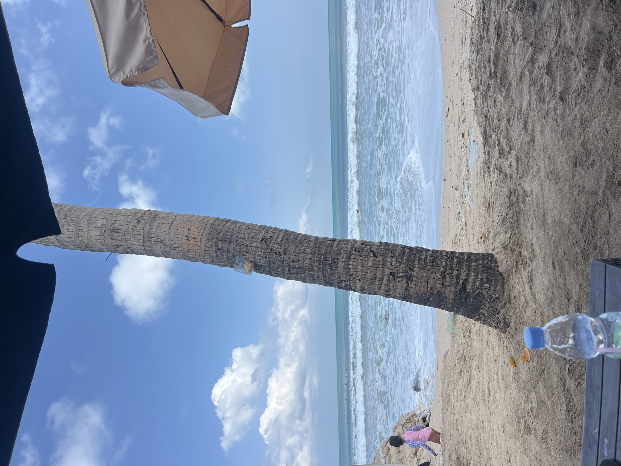 The image depicts a beach scene in Kuta, Indonesia. In the foreground, a dark wooden table holds a clear plastic water bottle with a blue cap and a pink straw. To the left of the bottle, a black object, possibly a phone, is partially visible. The sand is a light brown color.
Further back, a large, textured palm tree trunk dominates the center of the frame. A clear plastic bag containing yellow objects is affixed to the tree. To the left of the palm tree, a young child wearing a pink and blue swimsuit is standing on the sand and appears to be walking towards the ocean. The child is facing away from the camera. Behind the child and the palm tree, the turquoise ocean stretches to the horizon, with white waves breaking on the shore. Several distant figures, possibly surfers, are visible in the water.
Above the ocean, the sky is a bright blue with fluffy white clouds. On the right side of the frame, a portion of a beige beach umbrella is visible, providing shade. The overall environment is a sunny beach with a tropical feel.