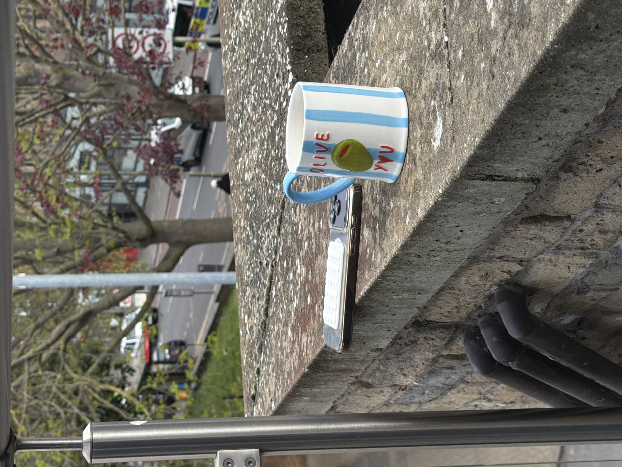 The image presents a close-up view of a white mug and a smartphone resting on a textured, grey concrete ledge or wall, overlooking a busy city street scene. The mug is decorated with light blue horizontal stripes and features the playful phrase "OLIVE YOU" in red capital letters, with a green olive illustration between the words. It has a light blue handle. The smartphone, which appears to be an iPhone, is positioned on its side, partially hidden behind the mug, and has a white, ribbed accessory attached to its back.
In the immediate foreground, a shiny metallic grey railing runs horizontally across the bottom of the frame, indicating the photograph was taken from an elevated vantage point, possibly a bridge or an upper floor. Beneath the concrete ledge, several dark pipes are visible against a roughly textured undersurface.
The background, although slightly out of focus, depicts an urban setting under bright daylight, suggested by clear shadows cast by objects. A street is visible with traffic, including cars and a white vehicle that resembles a police van with blue and red markings, typical of UK emergency services. Mature trees with both green and reddish-purple foliage line the street, partially obscuring distant buildings. No individuals are clearly identifiable in the scene. The bright conditions and urban environment are consistent with the context of London, United Kingdom.