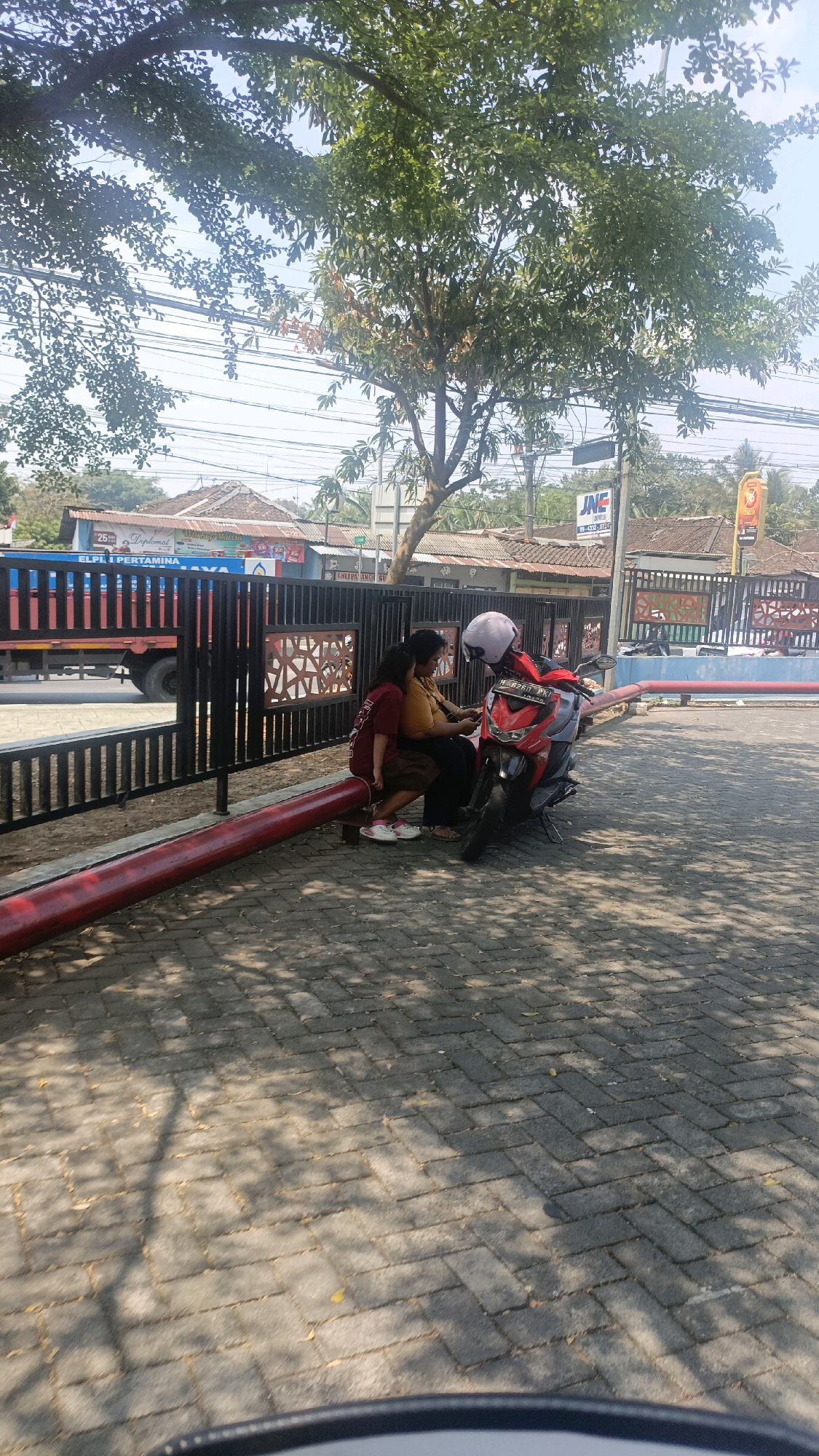 Two individuals are seated on a red metal beam in front of a black metal fence. One person, wearing a maroon shirt and brown pants, is seated on the left, leaning slightly towards the other person. The second individual, wearing a mustard-colored shirt and dark pants, is seated to the right, looking down at something in their hands. Between them is a red motorcycle with a pink helmet on the handlebars. The background shows a street with a red and blue truck, buildings with tiled roofs, and utility poles with wires. The ground is paved with interlocking gray bricks, and the scene is dappled with sunlight and shadows from overhead trees. A sign with "JNE EXPRESS" is visible in the distance. The location is identified as Salatiga, Indonesia.