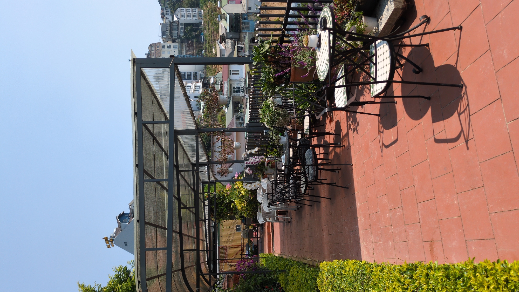 The image shows an outdoor patio or terrace in Da Lat, Vietnam. The space is covered by a metal-framed structure with a translucent roof. The flooring is made of terracotta-colored tiles. Tables and chairs are arranged along the patio, with potted plants and flowers decorating the space. In the background, there's a view of the city, with buildings scattered on the hillside. The sky is clear and blue. The plants and flowers contribute to a green and vibrant atmosphere. The overall scene suggests a relaxed and welcoming environment.