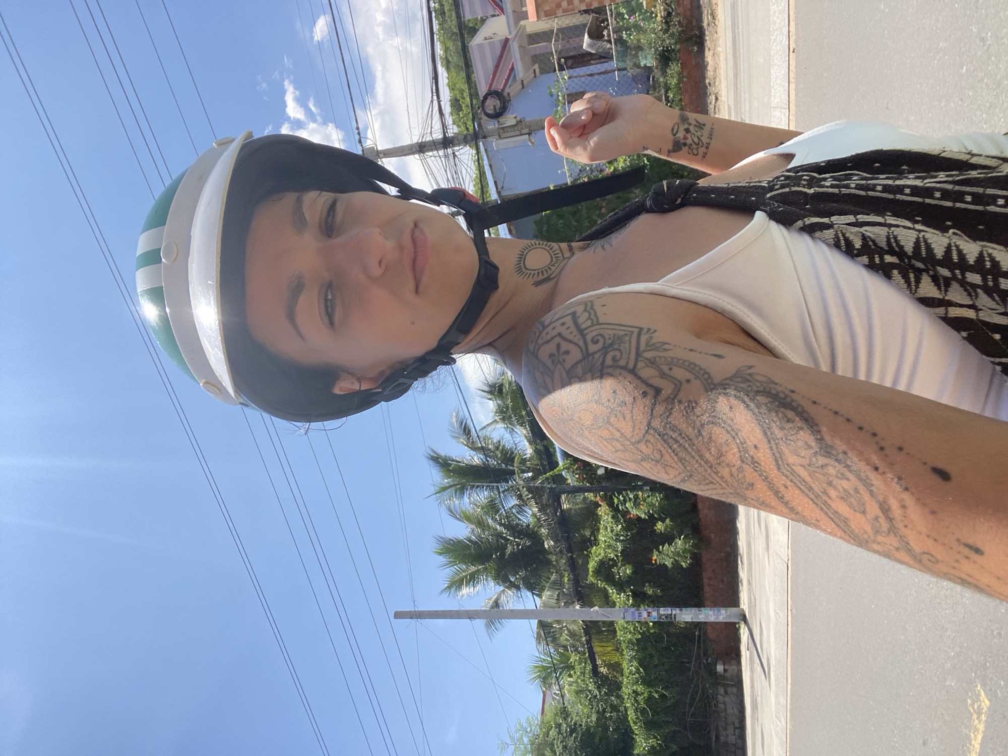A woman wearing a white tank top and a green and white helmet is taking a selfie while riding a scooter on a sunny day. She has a large tattoo on her left arm and a smaller tattoo on her right wrist with the initials "E.G.M." and a date. She is carrying a patterned bag over her shoulder. The background shows a street with power lines, palm trees, lush green foliage, and buildings. This scene is consistent with the environment found in Phu Thuy Ward, Vietnam, which is known for its tropical climate and vibrant street life.