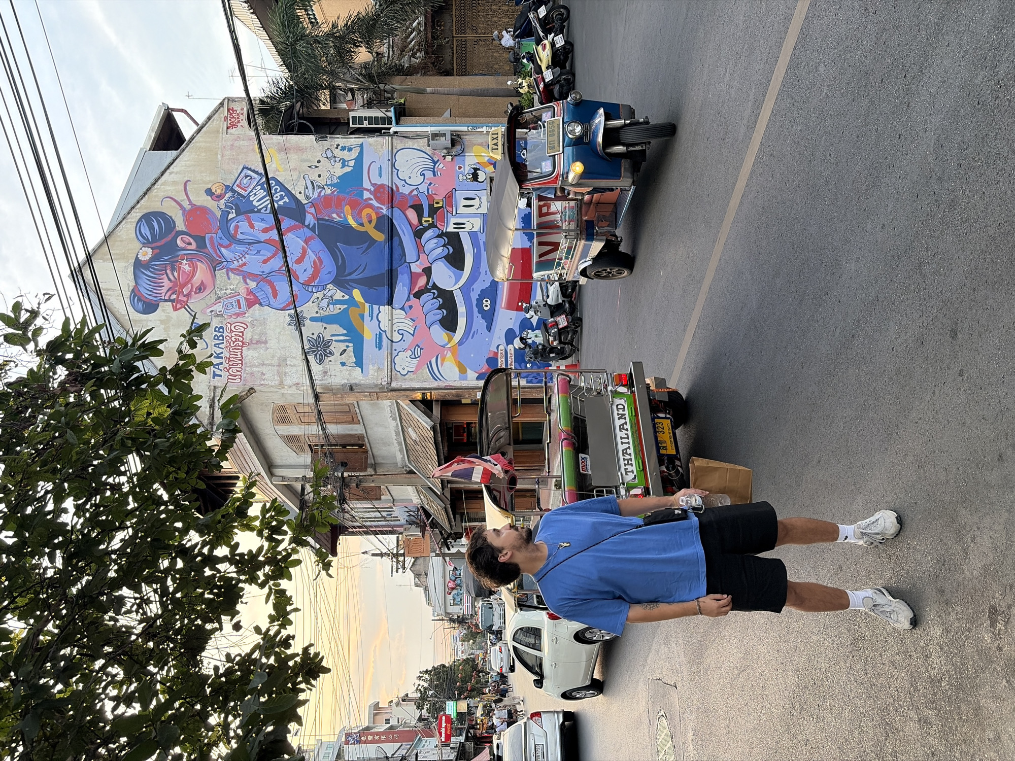 The photograph captures a street scene in what is likely Bangkok, Thailand. A man stands in the foreground on a paved street, dressed in a blue T-shirt and black shorts, with white socks and sneakers. He appears to be holding something in his right hand and has a shoulder bag.
The street is flanked by buildings, one of which features a large, vibrant mural on its side, depicting a cartoonish female figure. Two auto rickshaws (tuk-tuks), a common mode of transport in Thailand, are parked along the street. Several other parked motorcycles are visible as well.
Traffic is evident, with various cars and pedestrians further down the street, suggesting a busy urban setting. Above, a web of utility lines stretches across the sky. The lighting suggests it's either early morning or late afternoon, casting long shadows. A small amount of greenery is visible to the left.