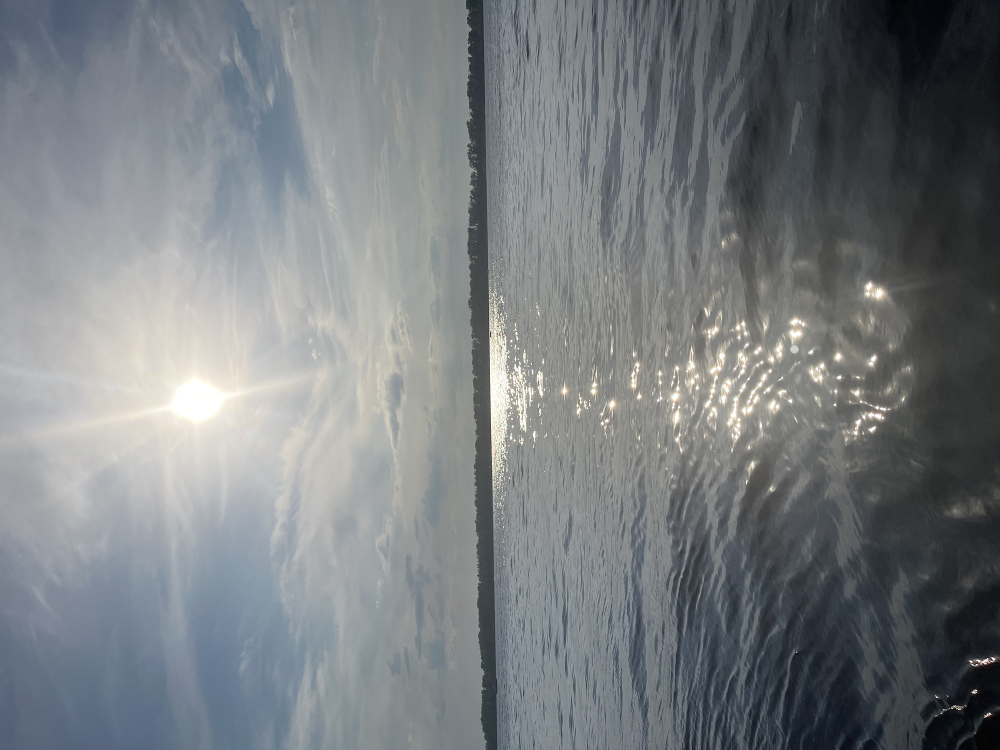 The image depicts a wide body of water with a treeline visible in the distance. The sun is high in the sky, partially obscured by wispy clouds, and its rays are reflecting brightly off the rippling surface of the water. The overall scene suggests a natural environment, likely a lake or a large river. Without further context or specific landmarks, it is not possible to definitively pinpoint the location within Novokuybyshevsk, Russia, but it is consistent with the presence of water bodies that can be found in or around the city. There are no people or specific man-made objects clearly visible in the scene.