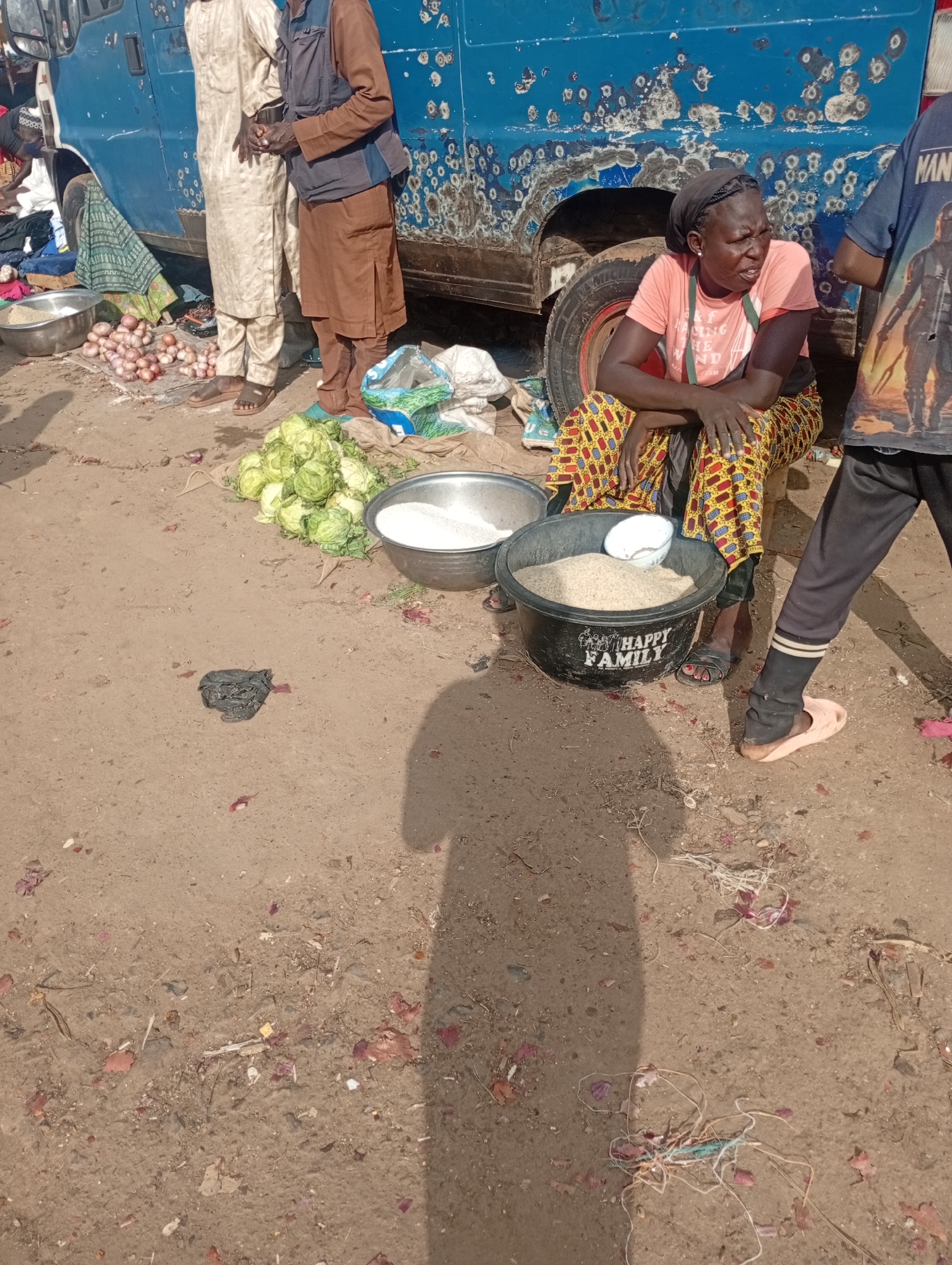 The image depicts an outdoor scene, likely a market, in Rafin Par, Nigeria. In the foreground, a woman is seated on a low stool, wearing a pink t-shirt and a colorful patterned skirt. She has a dark headscarf and appears to be looking towards her left with a neutral expression. Beside her is a large black bucket filled with white grains, possibly rice, and a smaller white bowl placed inside.
To the left of the woman, a pile of cabbages is visible on a woven mat, along with several red onions scattered on the ground. Two men are standing near a blue vehicle parked behind the produce. One man wears a light-colored traditional outfit, while the other has on a dark vest over a brown tunic and trousers.
The ground is dusty and unpaved, with scattered debris including pieces of red onion skins and bits of white and red string or plastic. The blue vehicle appears weathered, with rust and patches of paint missing. A shadow is cast on the ground from the right side of the frame, suggesting the sun is present, likely indicating daytime. The overall atmosphere appears to be a typical bustling market environment. The text "HAPPY FAMILY" is visible on the black bucket.