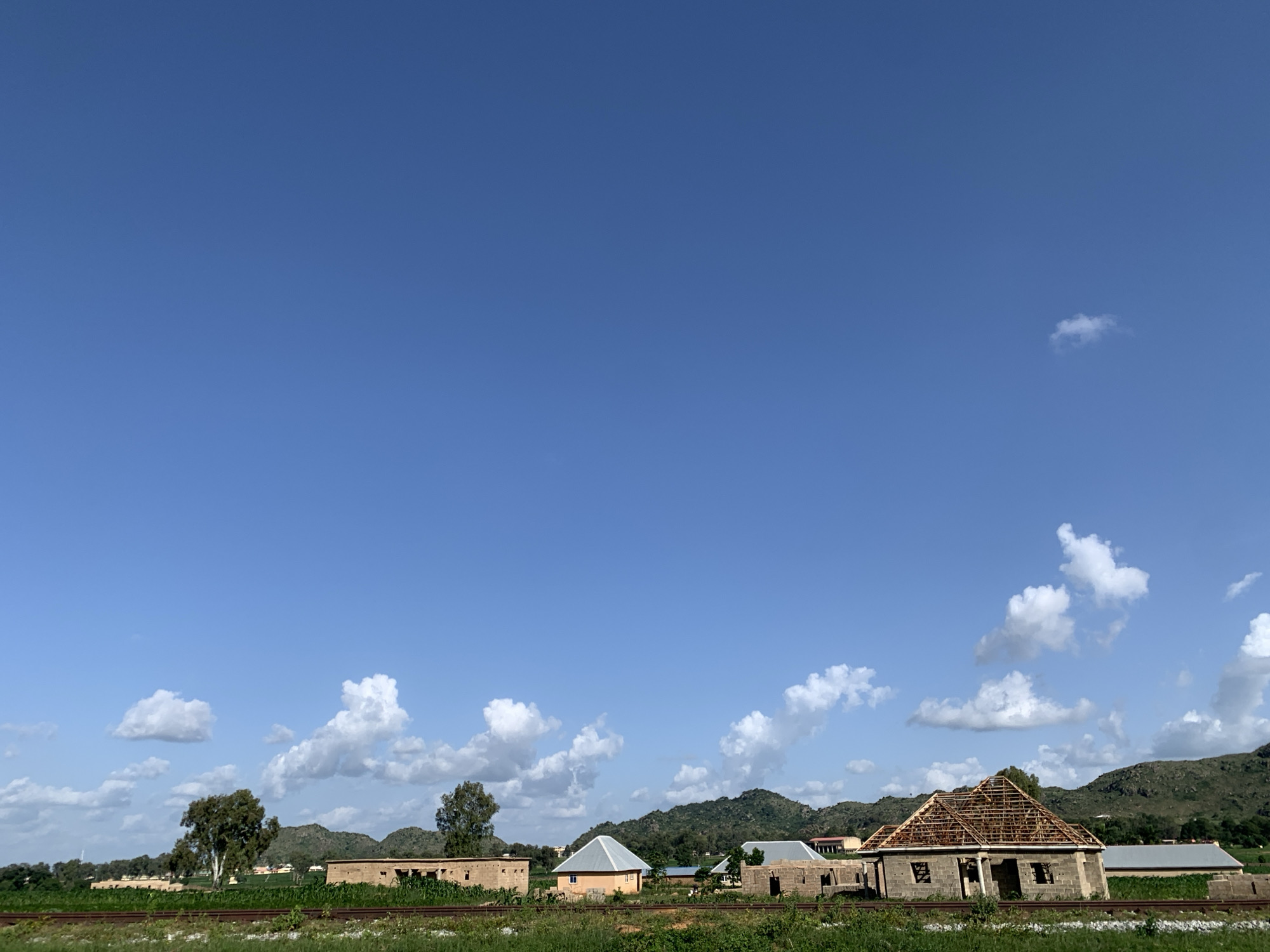 A wide shot captures a rural scene in Kuru Karama, Nigeria, under a clear blue sky dotted with fluffy white clouds. The foreground features a railroad track running horizontally across the frame, bordered by green grass and vegetation. Beyond the tracks, several unfinished and completed structures are visible, suggesting a developing area.
To the left, a long, low building with exposed brickwork stands partially constructed. Further to the right, a small, light orange house with a gray, pyramid-shaped roof is completed. Next to it, another building is under construction, with exposed wooden roof framing. Other buildings, some with intact roofs and others in various stages of completion, are scattered throughout the middle ground.
In the background, rolling green hills form a natural backdrop, with trees dotting their slopes. The overall impression is one of a quiet, rural community undergoing development. No people are visible in the image.