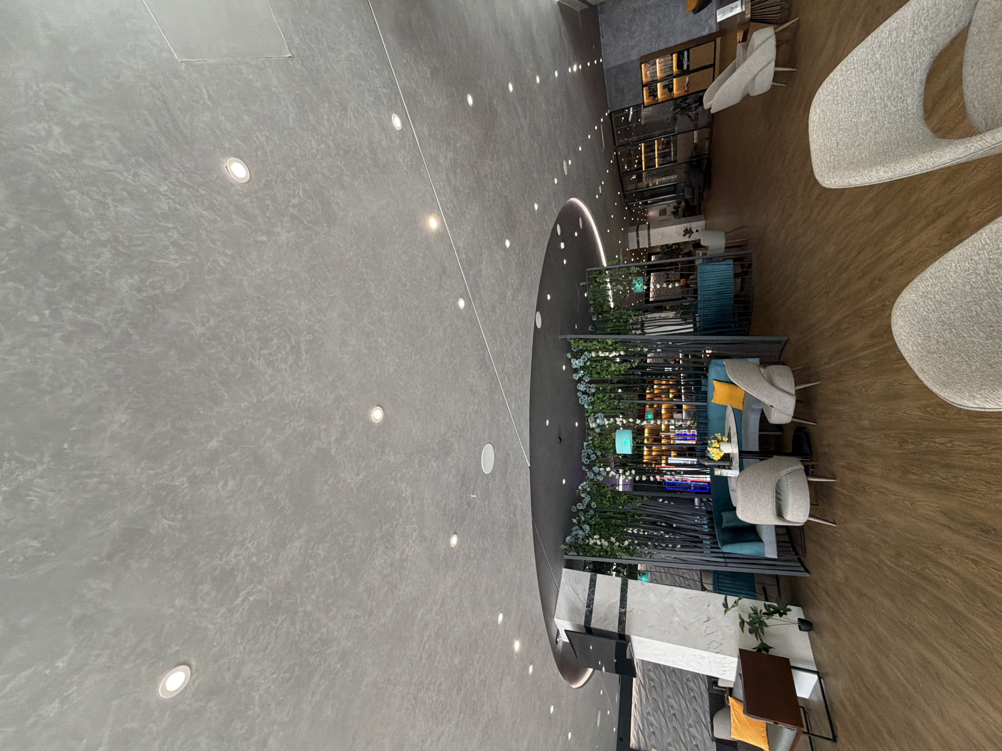 The image depicts a wide-angle view of a modern interior space, likely a cafe or lounge area, in Hisarya, Bulgaria. The scene is captured from a high vantage point, looking down and across the room. The flooring is a mix of textured grey material and polished wood.
The primary subjects are the seating arrangements. There are several comfortable-looking chairs with grey upholstered seats and backs, some with prominent curves. A few of these chairs are arranged around small tables. In the background, a bar or retail area is visible, featuring shelving units stocked with various items, illuminated by warm lighting. Decorative elements like artificial plants and flowers are strategically placed throughout the seating areas, adding a touch of greenery.
The lighting in the space appears to be a combination of recessed ceiling lights, which create a pattern across the grey ceiling, and warmer, more ambient lighting emanating from the background shelves and perhaps from within some of the decorative elements.
The time of day is not explicitly indicated, but the interior lighting suggests it could be evening or a dimly lit daytime environment. The overall atmosphere appears calm and inviting, with no people actively visible in the frame, suggesting a quiet moment or that the photo was taken before or after a busy period. The modern design, clean lines, and neutral color palette contribute to a sophisticated and contemporary feel. The location cues are limited to the provided context of Hisarya, Bulgaria. No visible text is present in the image.