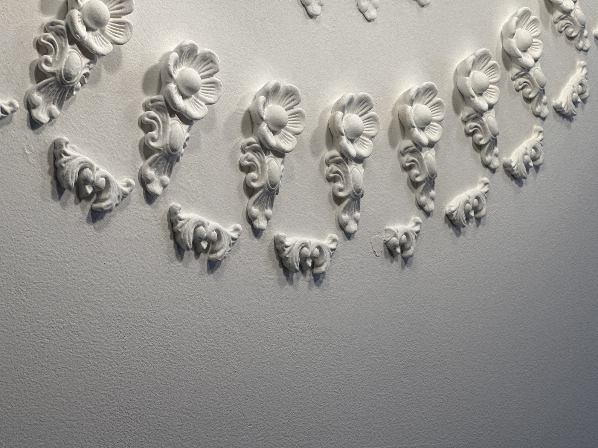 A detailed, high-angle shot showcases a textured white wall adorned with a series of ornate white plaster embellishments. These decorative elements, varying in size and design, are arranged in a diagonal line running from the top right to the bottom left of the frame. They feature floral motifs and scrolling acanthus leaves, characteristic of classical architectural detailing. The lighting creates distinct shadows, accentuating the three-dimensional quality of the reliefs and the rough texture of the wall. The image appears to be an interior detail shot, possibly from a historic building or a room with decorative plasterwork. The location is identified as Yessentuki, Russia, implying the context of a building within that city.