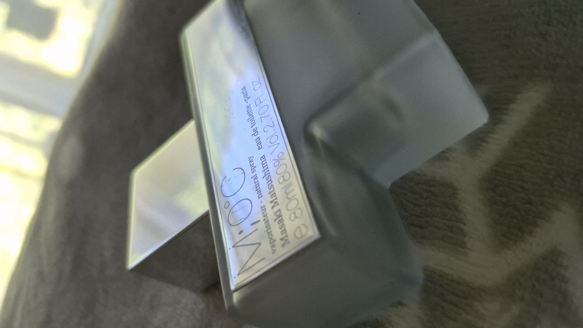 The image shows a close-up, slightly angled shot of a bottle of Masaki Matsushima M.o.c perfume. The bottle is made of frosted glass and has a metallic label. Text on the label reads "M.o.c", "vaporisateur - natural spray", "Masaki Matsushima", "eau de toilette - paris", and "e 80ml 80% Vol. 2.70 Fl. OZ.". The bottle is resting on a textured, fuzzy grey surface, possibly a blanket or fabric. In the background, a blurred, out-of-focus area suggests a bright, possibly natural light source, creating a soft glow. There is a metallic cube-shaped object to the left of the perfume bottle. The scene is indoors. No specific location in Gdańsk, Poland can be determined from the image.