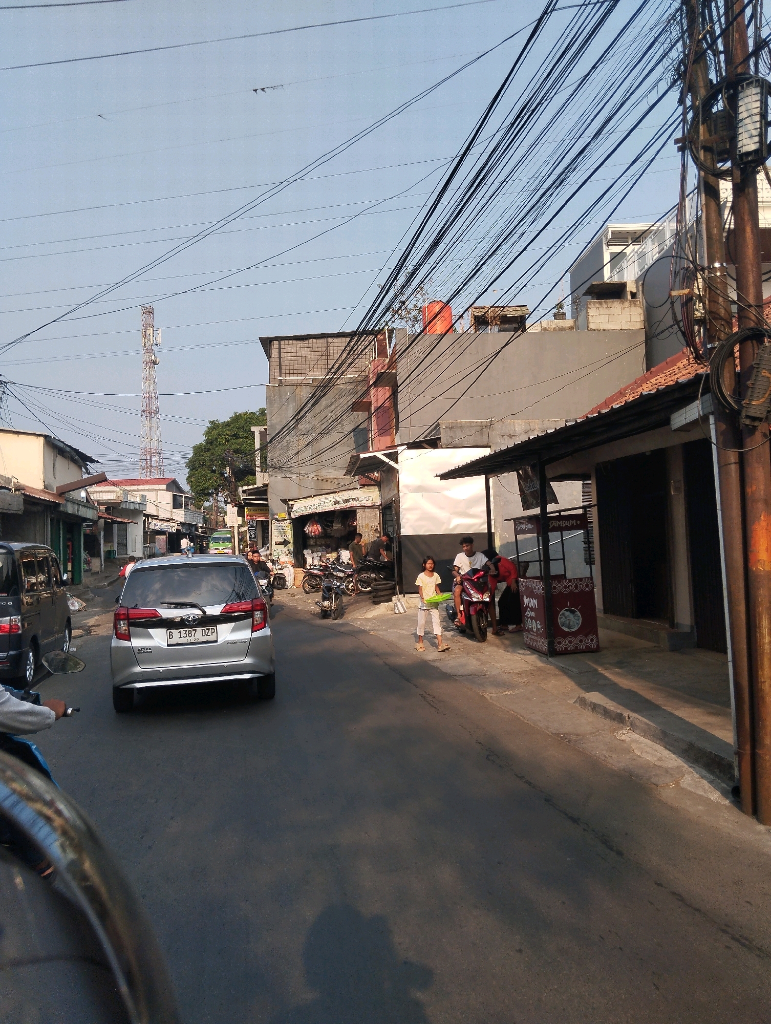 The image depicts a street scene in Sukaraja, Indonesia. In the foreground, a silver car with license plate B 1387 DZP is prominently visible. To the left of the car, a black van is parked. A person is riding a motorcycle in the lower left corner of the frame, with the motorcycle's handlebars and a portion of the rider's arm visible. The street is paved and appears to be a typical urban environment with buildings lining both sides. On the right side of the street, there is a small food stall with a red and white patterned advertisement. Several people are present, including a young girl holding a green tray and a young man on a red motorcycle. Further down the street, a transmission tower is visible in the distance, indicating the presence of telecommunications infrastructure. Overhead, a complex network of electrical wires crisscrosses the sky. The overall scene suggests a busy commercial or residential street with active daily life.