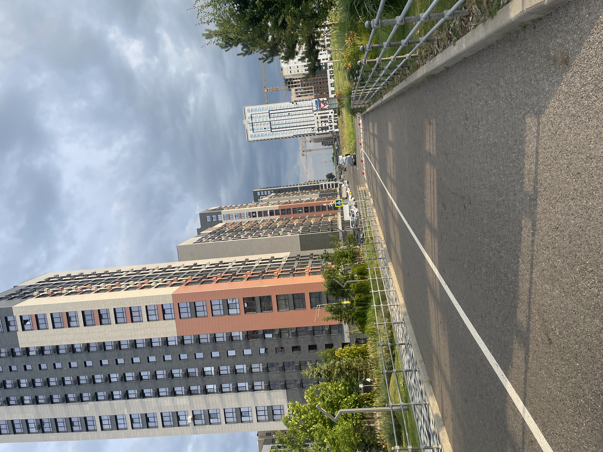 The image depicts an outdoor scene on a cloudy day, likely in a modern urban development area in Stavropol, Russia. A paved road with a white dividing line runs through the center of the frame. Along the sides of the road are metal railings and landscaped areas with grass, trees, and bushes. In the background, several tall, contemporary apartment buildings are visible. Some of these buildings have a mix of grey and light-colored facades, with numerous windows. One building features an orange accent. In the distance, construction cranes indicate ongoing development. Several vehicles, including cars and what appear to be utility vehicles, are present on the road and parked nearby. The overall impression is one of a newly developed residential district.