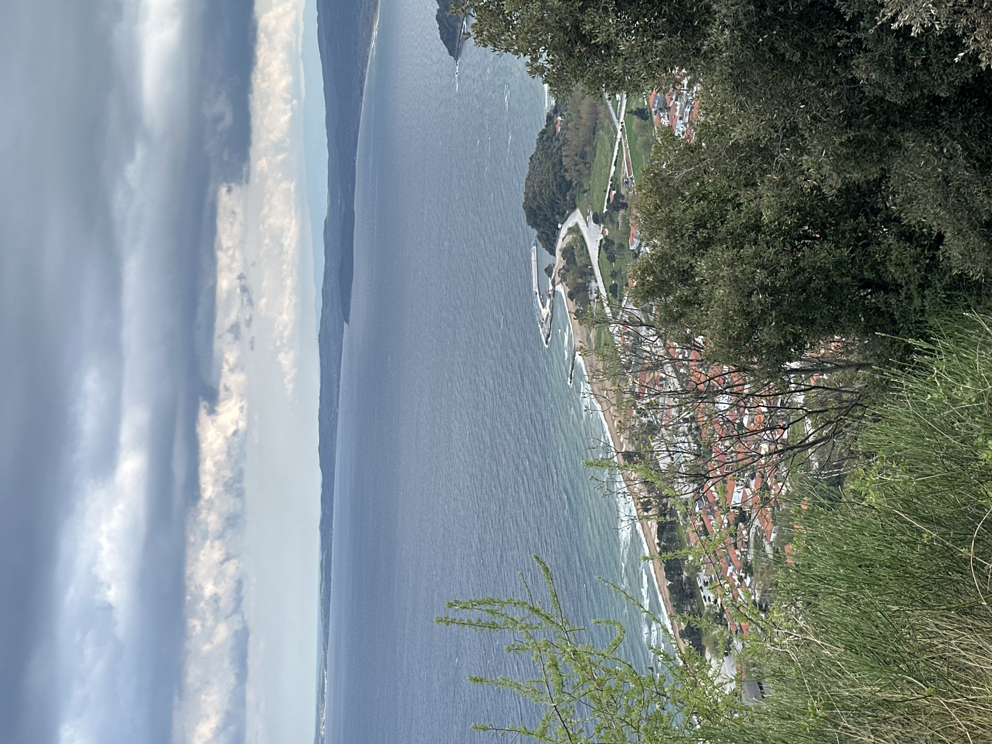 An elevated landscape view, shot in portrait orientation, overlooking the coastline of Stratoni, Greece. The Aegean Sea stretches from the mid-left to the top-left of the frame, bound by a distant landmass. The city of Stratoni runs along the coast on the right, with rooftops and infrastructure visible. The vegetation of the area surrounding the city is on the right side. Weather conditions are partially cloudy.