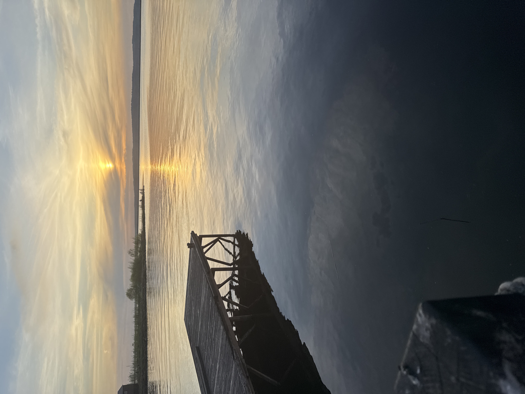 A view of the Kama River in Chaikovsky, Russia. The sun is setting on the horizon, casting yellow and orange colors on the water. A pier juts out into the river. The water reflects the sky. The sky is a mix of clouds and blue. A small piece of land is visible on the horizon. A building is located to the bottom left of the pier.