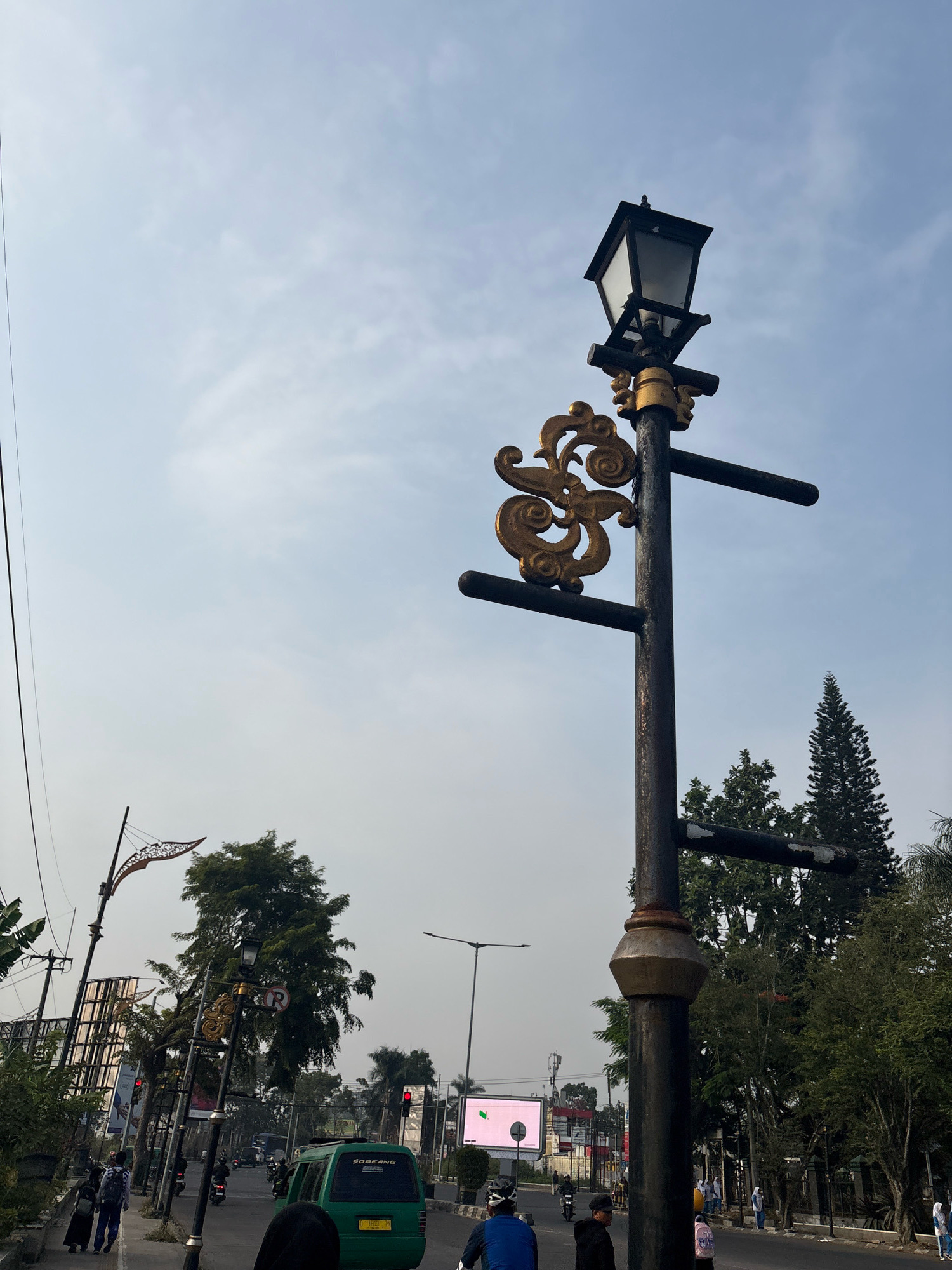 The image depicts a street scene in Pamekaran, Indonesia, under a cloudy sky. In the foreground, a decorative lamppost with ornate gold scrollwork stands prominently. To the left, a sidewalk is populated by several pedestrians, including two individuals walking away from the camera, one carrying a backpack. A green van is visible on the road, along with several motorcycles and other vehicles further down the street. Traffic lights can be seen in the distance. Trees line the street, some of which are tall and conical. A large billboard is also present on the left side of the image. The overall impression is of a busy urban environment.
