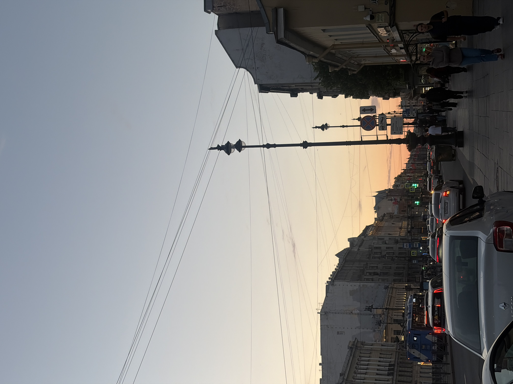 The image depicts a street scene in Saint Petersburg, Russia, during what appears to be twilight. The sky is a gradient of pale blue and soft orange, indicating the sun has recently set or is about to rise. Multiple power lines crisscross the sky. On the right side of the street, a row of buildings with detailed architecture lines the sidewalk. The buildings vary in color and style, with some featuring ornate facades and others appearing more utilitarian. One building has a yellow facade with storefronts. On the left side of the street, more buildings of similar architectural style are visible.
The street is busy with traffic. Several cars are stopped, suggesting a traffic jam or intersection. A blue tram with the number '42' displayed on its front is visible on the far left. Pedestrians are walking along the sidewalk on the right side of the street, some in groups and others individually. A tall, ornate lamppost stands prominently on the sidewalk, casting a shadow. Several street signs are also visible, including a circular sign with a red border and a diagonal red bar, and a sign with an arrow indicating direction. The pavement is paved with cobblestones. The overall atmosphere is that of a bustling city street at dusk.