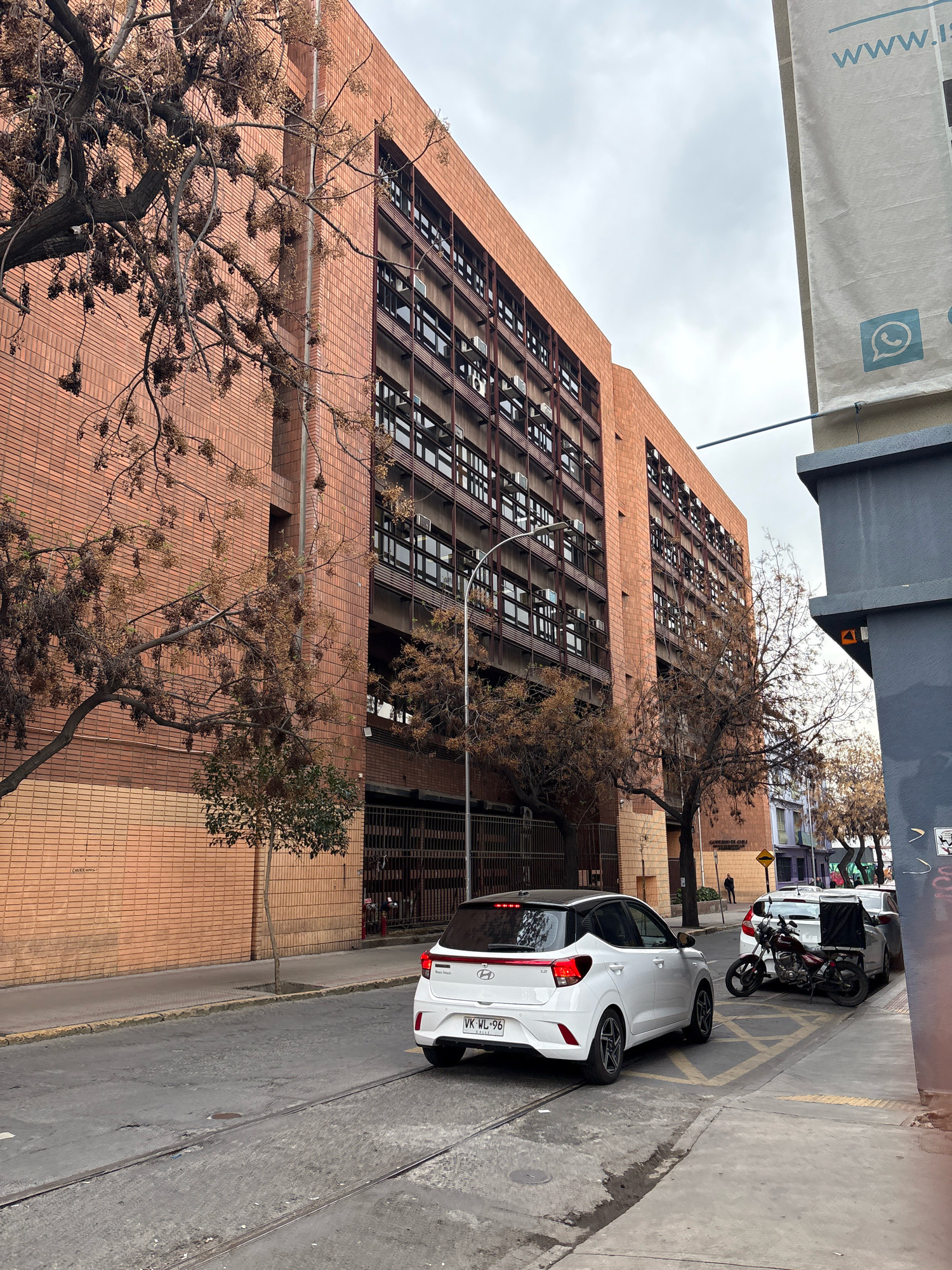 This image depicts a street scene in Santiago, Chile, on an overcast day. A multi-story building with a brick facade and numerous windows dominates the background. In the foreground, a white car with a black roof is parked on the right side of the street, its rear facing the camera. A motorcycle is parked next to it. A street lamp is visible on the left side of the image, and trees with bare branches line the sidewalk. The street itself appears to be paved, with tram tracks running through it. A large banner with text and a WhatsApp logo is affixed to a building on the right side of the frame. The overall impression is one of an urban environment with a muted color palette due to the weather conditions.