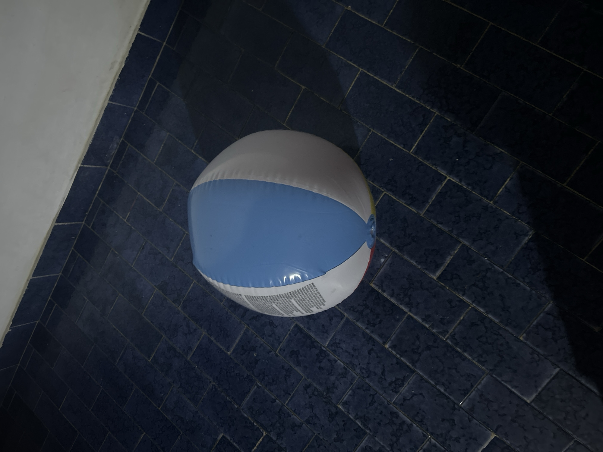 A slightly deflated, multi-colored beach ball rests on a dark blue tiled floor. The tiles appear to be arranged in a herringbone pattern. The wall is visible on the left edge of the scene. It's a sterile, indoor environment. Location unknown.