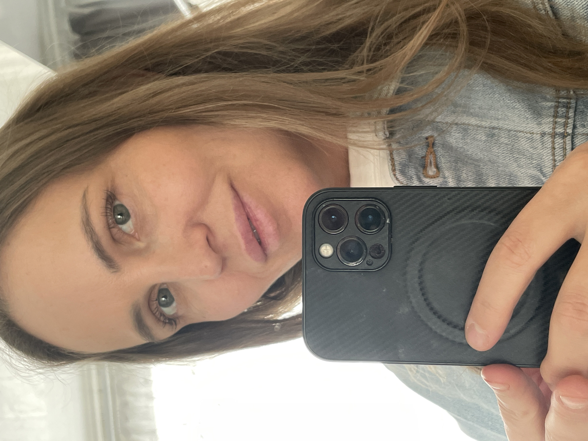 A female individual is depicted in a mirror selfie, presenting a close-up view from the chest up. She has long, light brown hair, light skin, and green eyes, with subtle makeup. A faint smile is visible on her face. She is wearing a light-wash denim jacket over a white top. In the foreground, she holds a black smartphone with her left hand, obscuring part of her body. The phone features a prominent triple-lens camera module, a flash, and is encased in a textured black cover with a visible circular MagSafe ring impression. The background is overexposed and largely white, suggesting a brightly lit interior space, possibly near a window, with faint reflections of what appears to be a curtain or light-colored wall visible on the upper right. The specific location within the city of Chaikovsky, Russia, is not discernible from the visual evidence in the image.