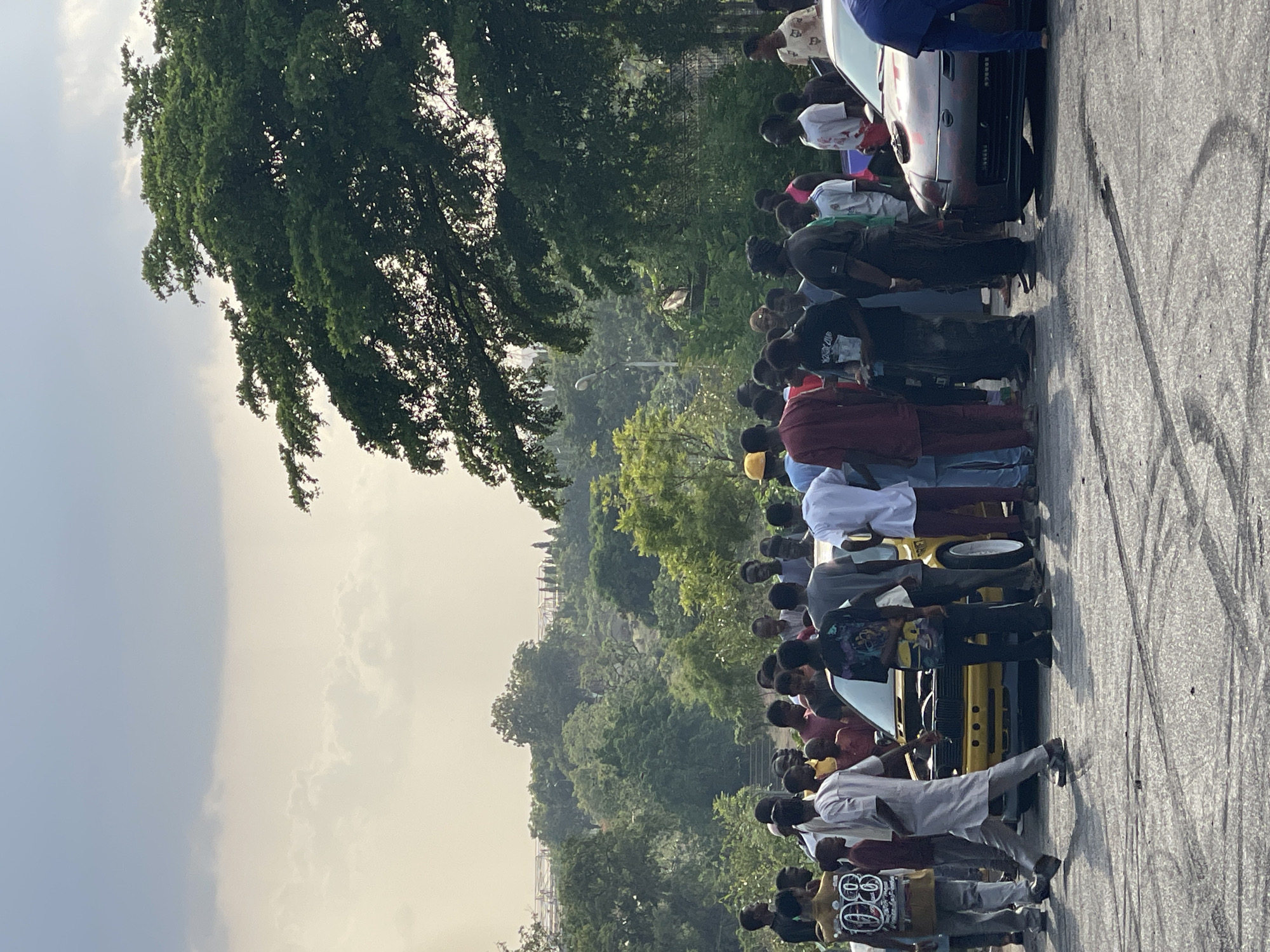 A large group of people are standing adjacent to cars, on what appears to be a stretch of road with skid marks. The location is likely in Abuja, Nigeria, indicated by the environment and context. The group is predominantly young people. Several cars of different colors are parked along the roadside. Trees and a cloudy sky can be seen in the background. The road has distinct skid marks, suggesting it may be used for racing or other car stunts.