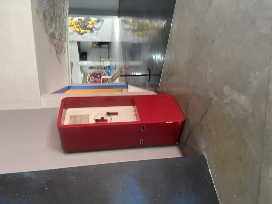 The image shows a red-colored, rectangular metal box with a white interior. It is located in an indoor space with gray concrete floors. There is text on the white interior, but it is too far to read. There is a blue wall to the left of the box and a white wall to the right, with a doorway leading to another area. The doorway has a blue, yellow and orange stripe painted along the top and left side. The other area is brightly lit and has shiny, gray concrete floors. A sign is standing on a black metal stand in this other area. The image appears to have been taken inside an indoor attraction or museum in Padalarang, Indonesia (latitude: -6.852, longitude: 107.494).
