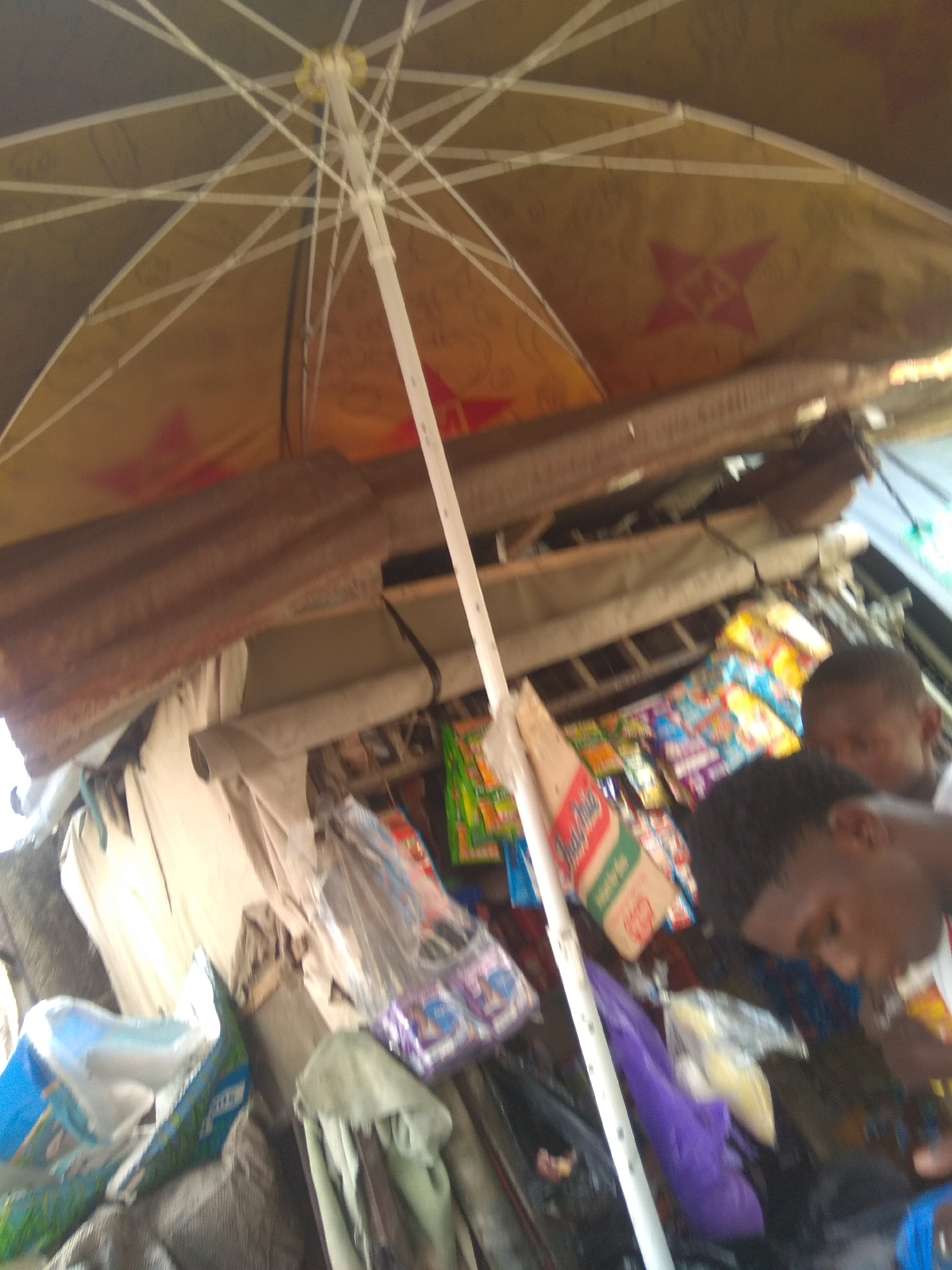 This media file depicts a street stall in Jalingo, Nigeria, likely during the daytime. The scene is dominated by an open umbrella, its yellow fabric patterned with red star shapes, providing shade over the stall. Below the umbrella, a makeshift structure is visible, possibly constructed from corrugated metal and wood, supporting an array of goods.
The stall is packed with various items, primarily packaged snacks and small goods. Brightly colored wrappers of snacks are piled high, some in plastic bags. A prominent red and green packaged item, possibly Indomie noodles, is visible. Among the merchandise, there are also purple and clear plastic bags containing items.
In the foreground, two young Black males are partially visible. One, in the lower right of the frame, is looking down and appears to be holding something in his hand. The other, further back and to his left, is also looking down. Their expressions are not clearly discernible due to the image quality and their positioning.
The overall impression is of a busy, albeit somewhat cluttered, small business operating outdoors. The lighting suggests it is daytime, and the presence of an umbrella hints at either bright sunshine or the possibility of light rain. The setting appears to be a public street or market area.