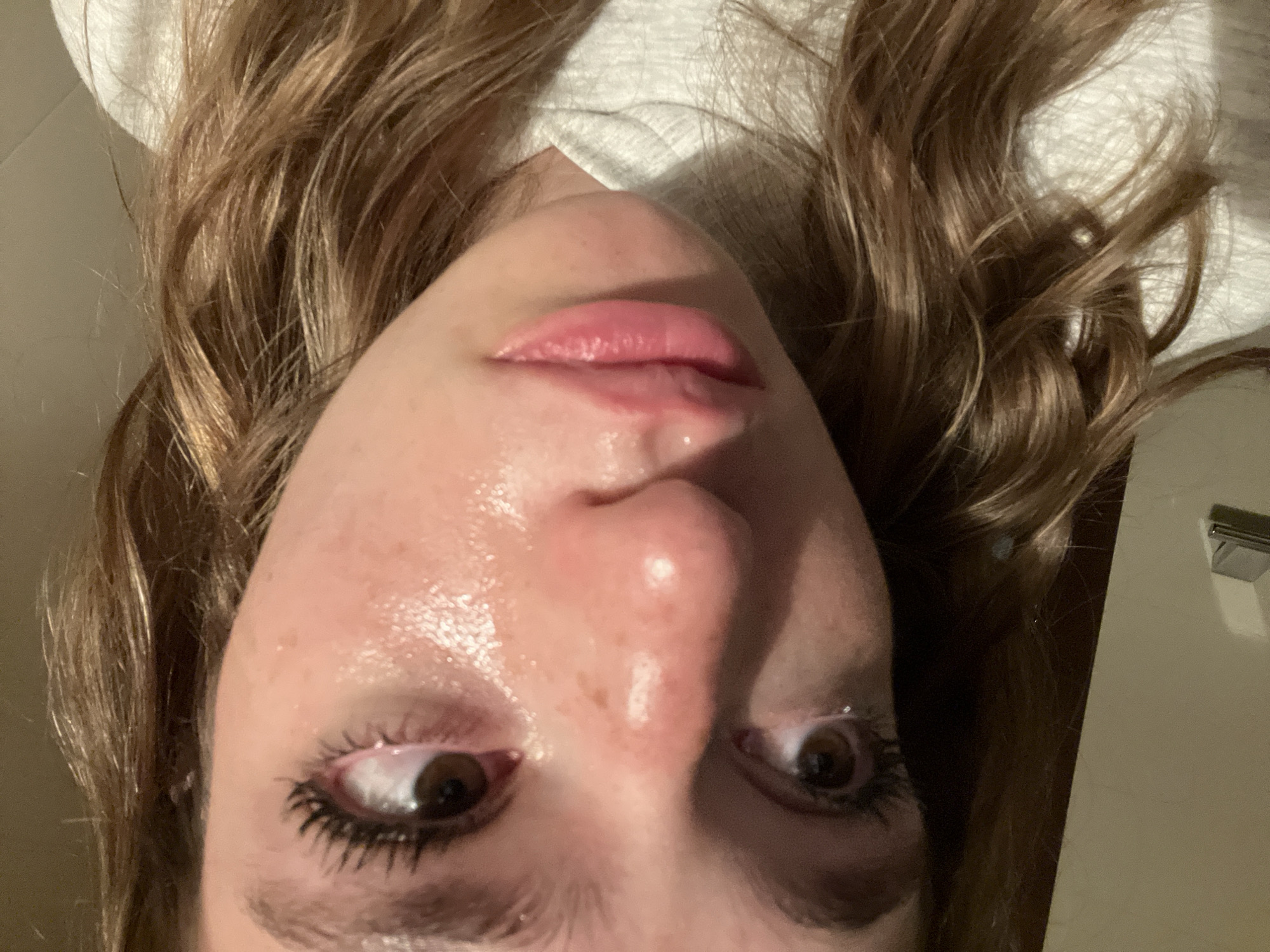 The image shows a close-up of a person's face from a low angle, looking upwards. The person has brown eyes with dark mascara, light skin with freckles, and full lips with a glossy sheen. Their hair is light brown and wavy, framing the face. The person is wearing a white shirt or top. The image appears to be taken indoors, with a light-colored wall visible in the background. The location is unknown.