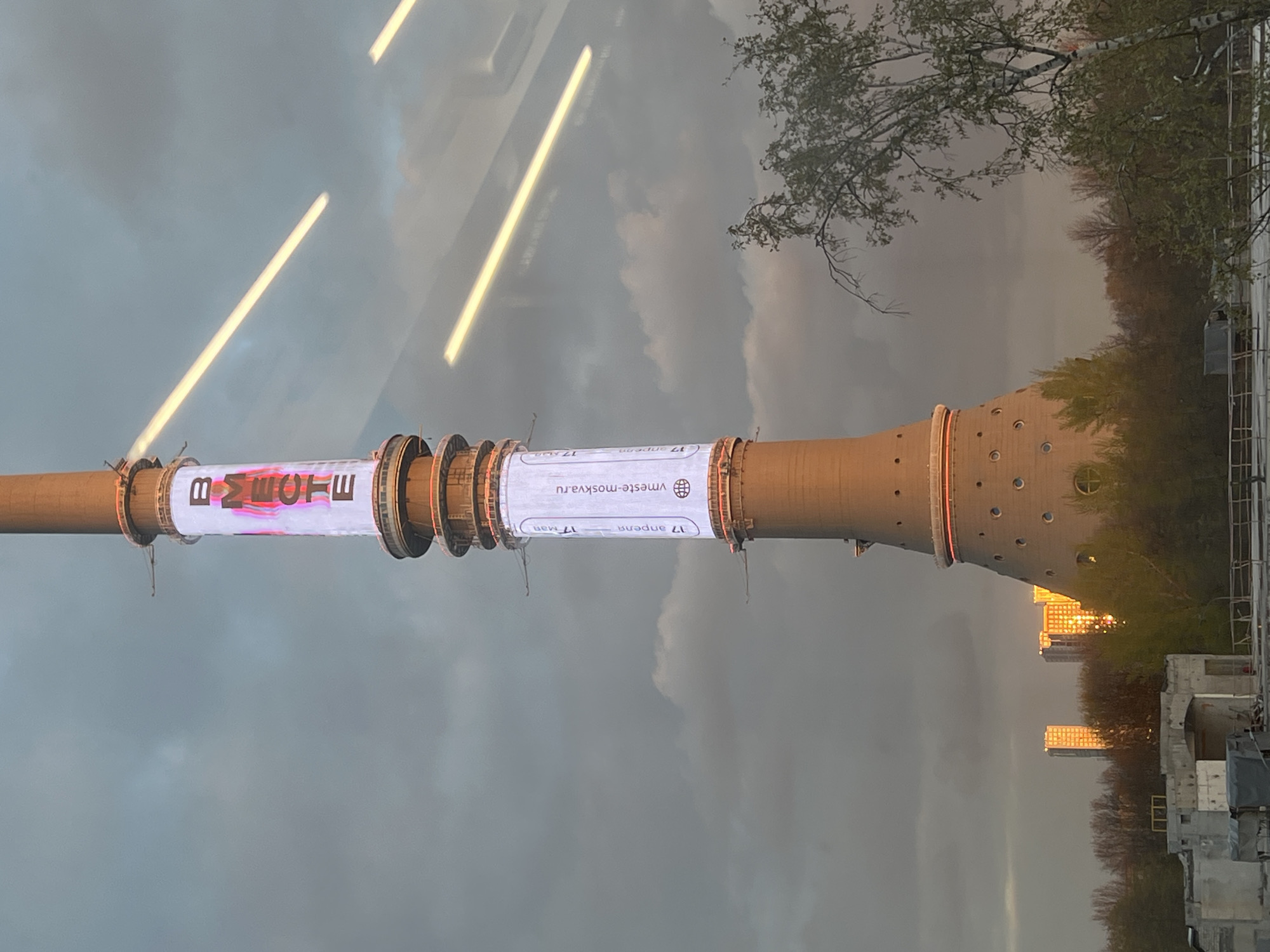A vertical outdoor photograph, likely taken through a window due to visible reflections in the upper left, depicts a tall, cylindrical structure, possibly a power plant chimney, against an overcast sky. The structure, made of light brown concrete, features several prominent rings or bands around its upper half and evenly spaced circular openings in its lower section.
A large banner is wrapped around the upper part of the tower. On its left side (when viewed upright), the Russian word "ВМЕСТЕ" (VMESTE), meaning "TOGETHER," is displayed in stylized, multi-colored letters with a dynamic, almost flowing texture. On the right side of the banner, a website address "vmeste-moskva.ru" is visible alongside a globe icon. Below this, two dates are printed: "17 апреля" (April 17) and "17 мая" (May 17), followed by a "Z" symbol or logo.
The sky is cloudy and grey. To the right of the tower, trees with sparse foliage suggest early spring. In the distance, towards the bottom right, several modern buildings reflect a strong, golden sunlight, indicating a specific time of day like late afternoon or early morning when the sun might be low or briefly breaking through the clouds. Some construction materials or scaffolding are visible at the bottom right. The overall setting is an urban or industrial area in Khimki, Russia.
