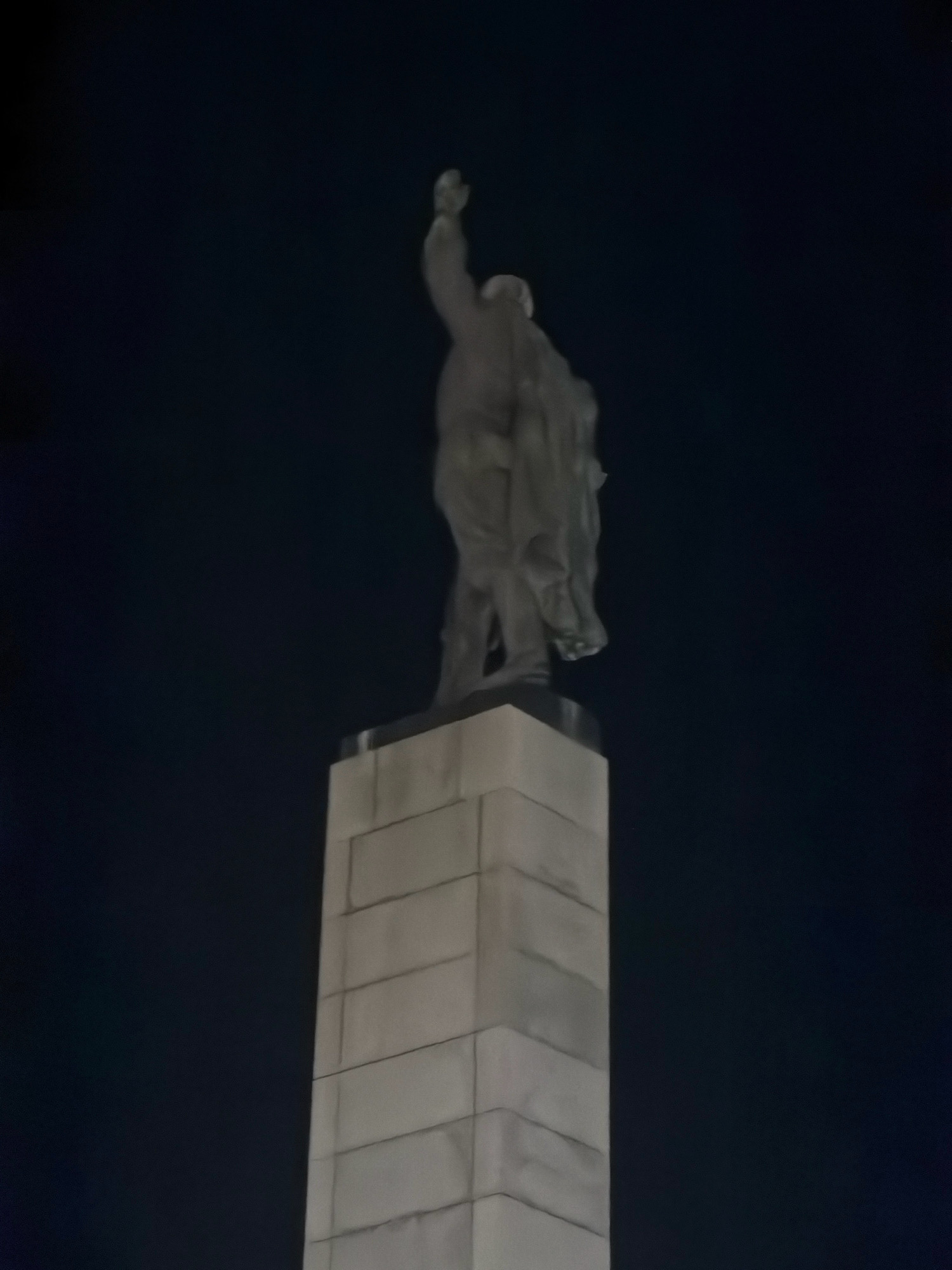 The image shows a statue of a man, likely a historical figure, raised on a tall, rectangular pedestal. The statue is made of a dark material and depicts the man with his right arm raised. The pedestal is constructed from light-colored stone blocks. The scene is illuminated from below, casting shadows and creating a dramatic effect against a dark night sky. The statue is located in the city of Burgas, Bulgaria.
