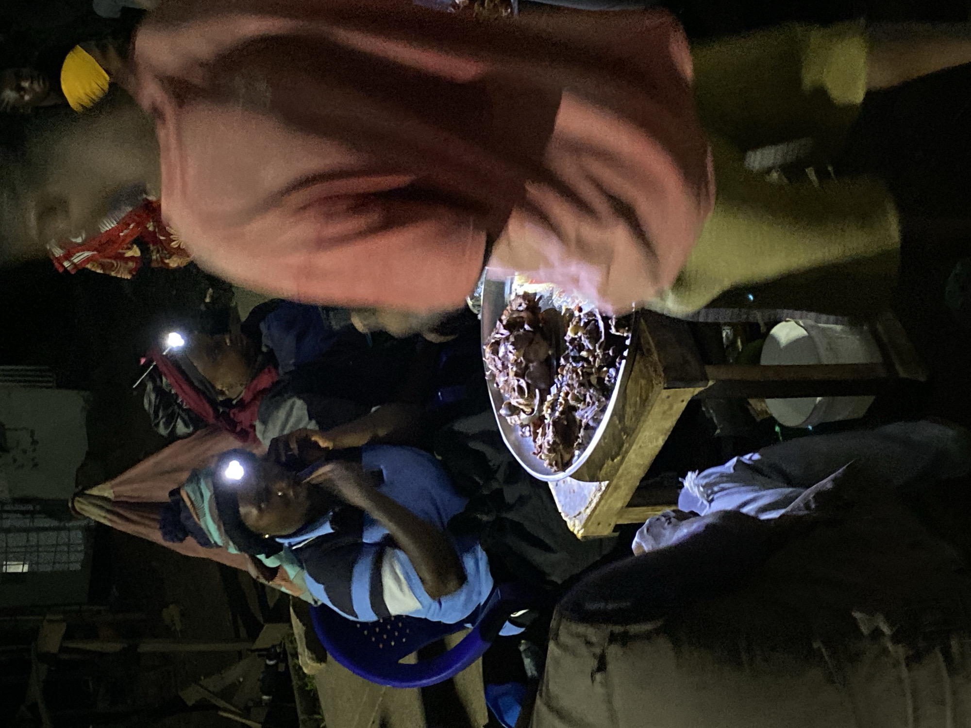 People are gathered around a table at night in Bukuru, Nigeria. The individuals are illuminated by headlamps. A large metal bowl on the table contains dark food items. People are eating. There is a blue plastic chair. The location appears to be outdoors. Other people are present in the background.