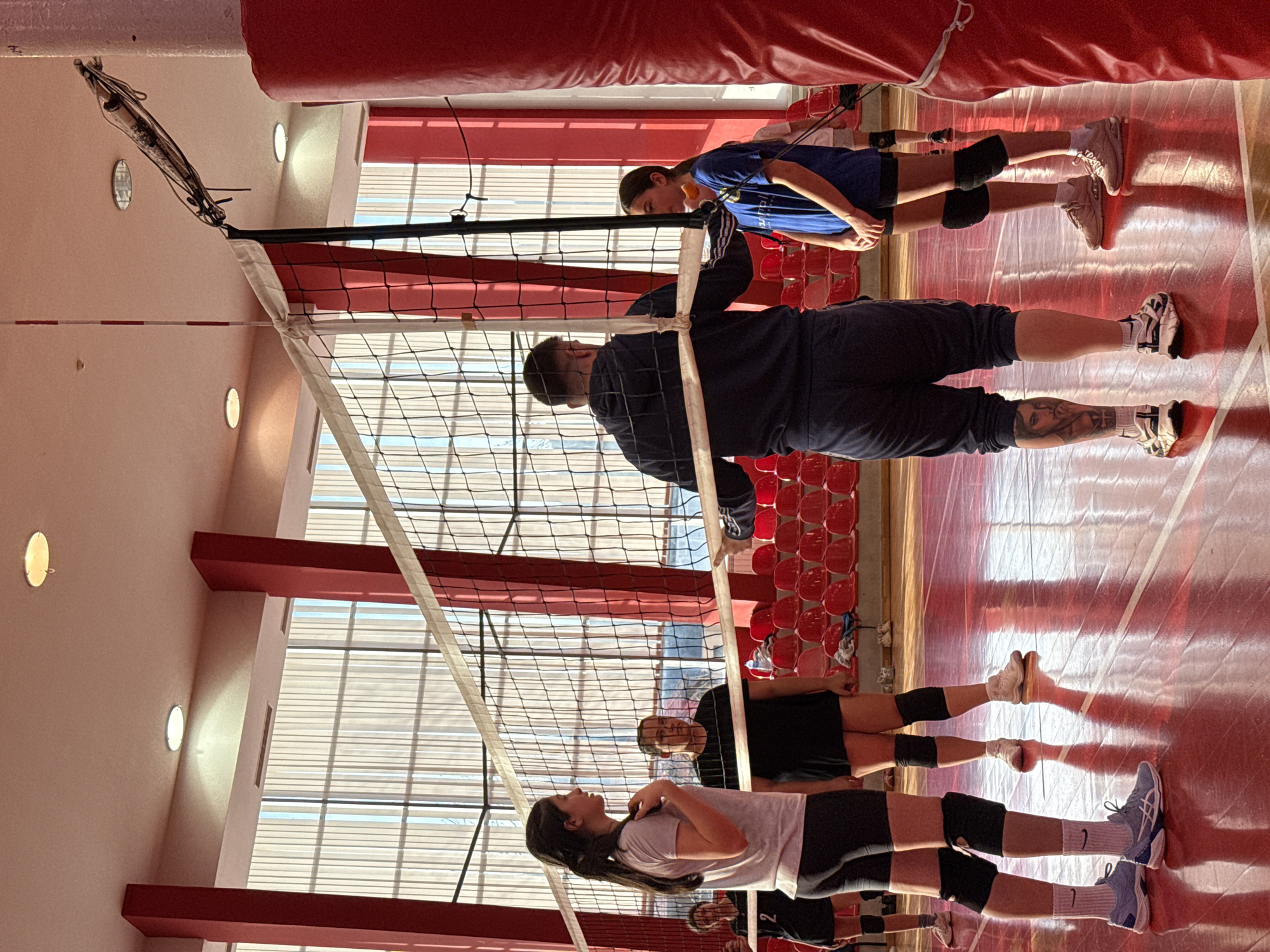 A volleyball net divides an indoor gymnasium. Several people are visible on either side of the net. On the left of the net, a young woman with long dark hair stands in a light-colored t-shirt and dark shorts, wearing knee pads and white socks, looking towards the right. Behind her is another person. On the right side, an adult male with a shaved head stands at the net wearing dark clothing and shorts, displaying tattoos on his legs. Behind him stands a younger person in a blue shirt and dark shorts, wearing knee pads. All are facing the net. The floor of the gymnasium is red with white lines. Red seating is visible in the background. Large windows provide light to the interior space. The gymnasium appears to be located in a building with red architectural features. The image does not contain explicit information about Etropole, Bulgaria.