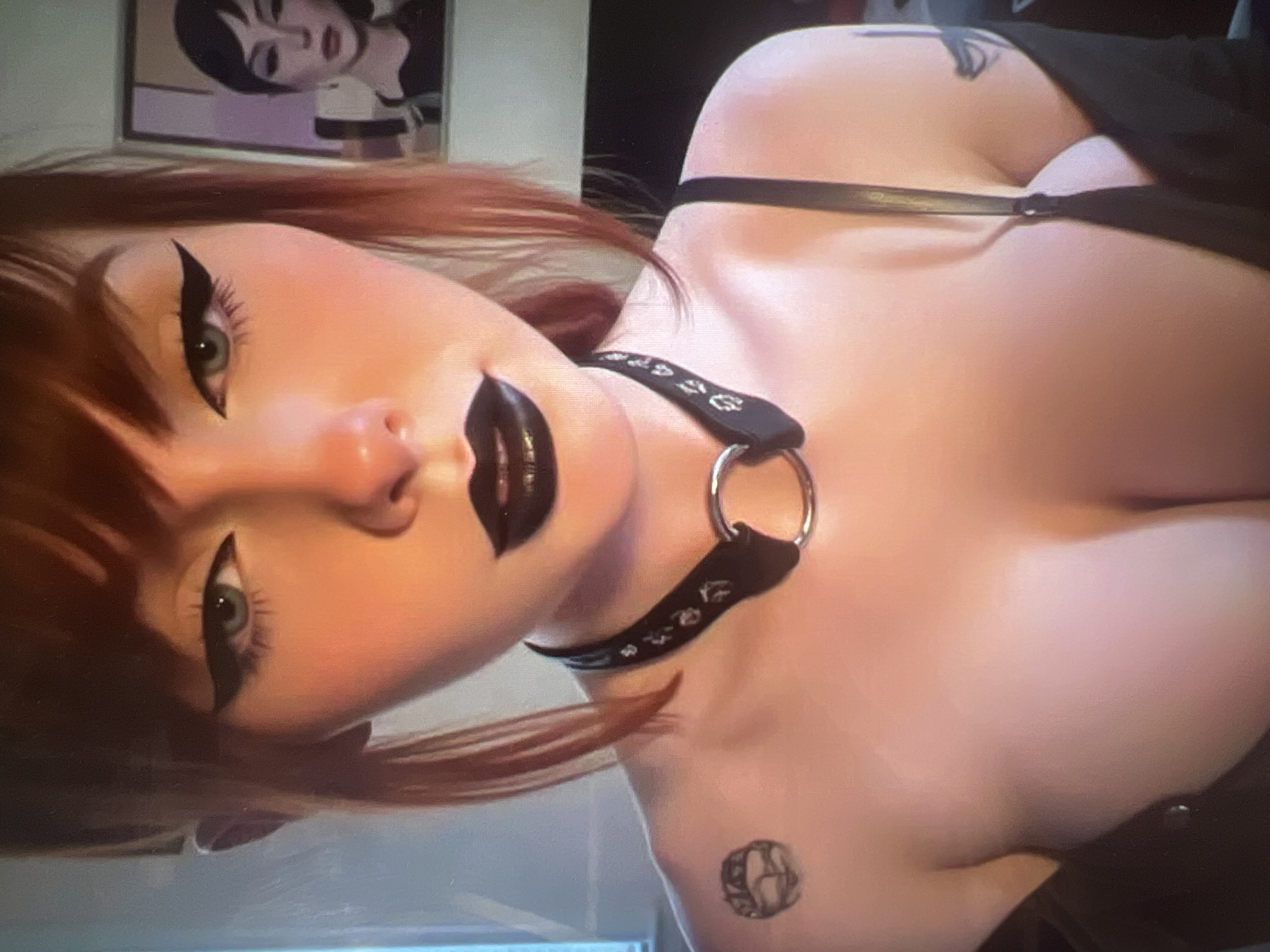 The image is a close-up, waist-up shot of a person with reddish-brown hair styled with bangs. They are wearing heavy black eyeliner and black lipstick. A black choker with a silver ring and decorative studs is around their neck. A tattoo is visible on their left shoulder. The person is wearing a black top with spaghetti straps, revealing their chest. Another tattoo is visible on their right shoulder. In the background, a framed image of a woman with dark hair and similar makeup can be seen. The lighting suggests an indoor setting. There is no discernible information in the image that can identify the location as Minsk, Belarus.
