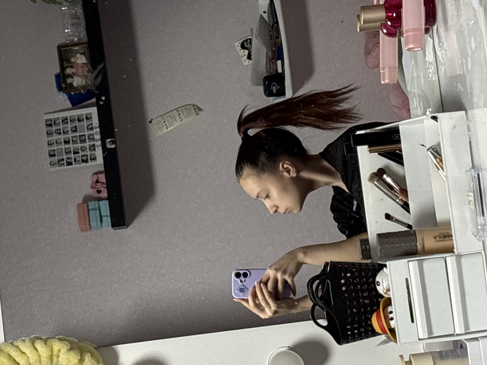 A young woman with dark hair in a high ponytail is standing in front of a vanity. She is holding a purple smartphone and appears to be taking a photo or video. The vanity is white and has various makeup items on it, including brushes, foundation, and pink lip glosses. A black purse is also visible on the vanity. The background is a light gray wall with some items hanging on it, including a framed photo and a calendar. The location is in Прикамский, Russia.