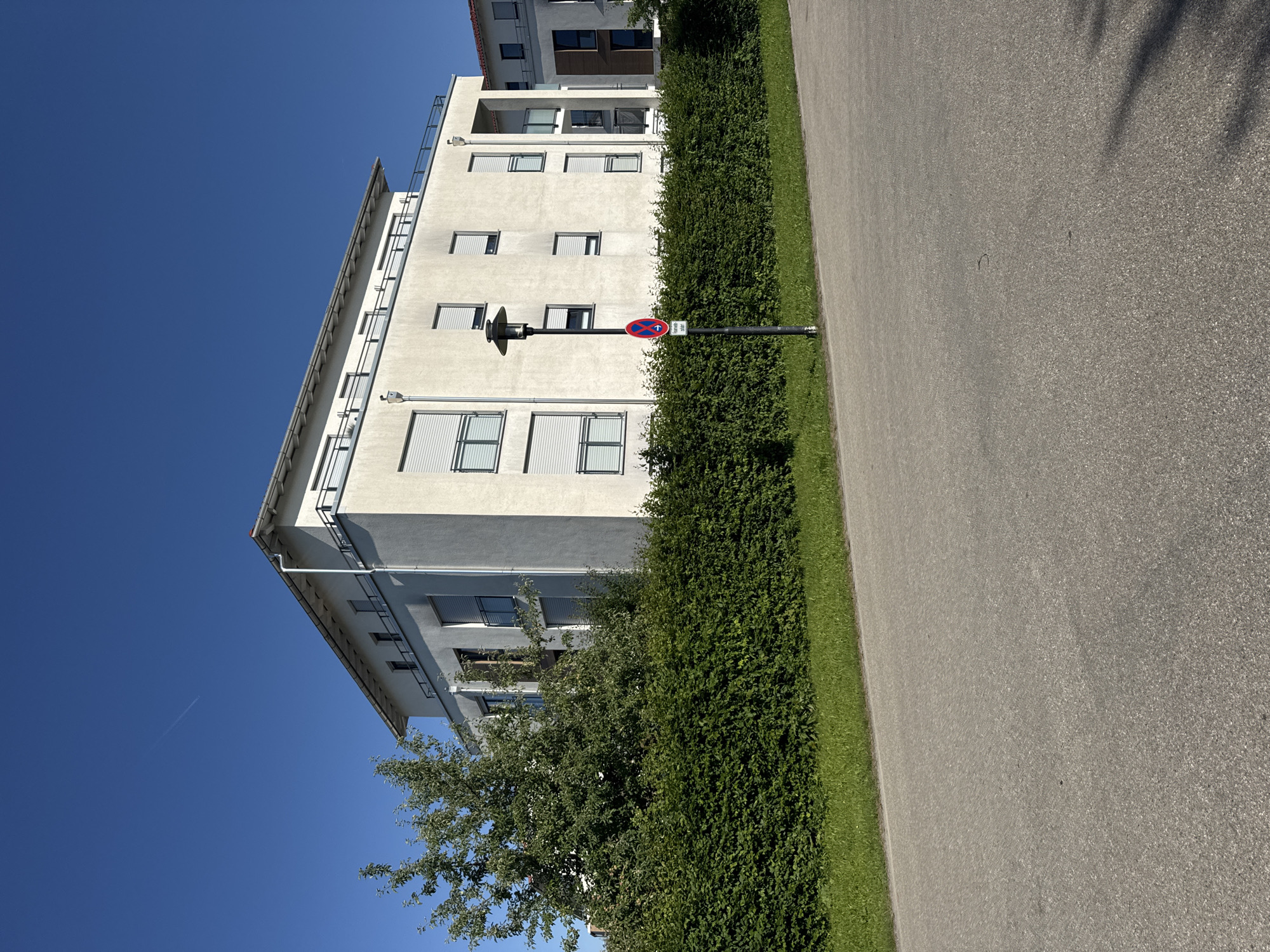 In Höhenkirchen-Siegertsbrunn, Germany, on a clear day, a multi-story modern apartment building with a light-colored facade stands behind a neatly trimmed green hedge. The building features several balconies with railings and windows with white roll-down shutters. A lamppost with a sign is positioned in front of the hedge. To the left of the building, a large green tree with dense foliage is visible. In the foreground, a paved pathway or road runs horizontally across the image. The sky is a vibrant, cloudless blue.