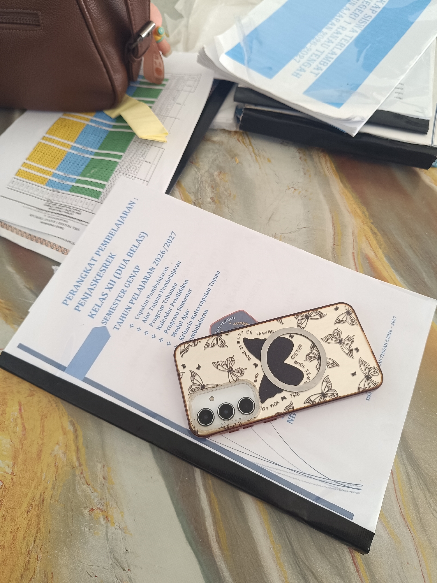 A top-down shot shows a variety of documents and a smartphone on a table. The documents are stacked and fanned out, with one document prominently displaying text in Indonesian, including "PERANGKAT PEMBELAJARAN", "PENJASKESREK", "KELAS XII (DUA BELAS)", and "SEMESTER GENAP". Another document has blue and white text, with some visible words in Indonesian. A brown leather bag is partially visible in the upper left corner. A smartphone with a butterfly-patterned case and a circular ring holder is placed on top of the documents. The background appears to be a textured surface, possibly a table. The scene appears to be indoors. It is not possible to determine the specific location in Jepara, Indonesia from the image alone.