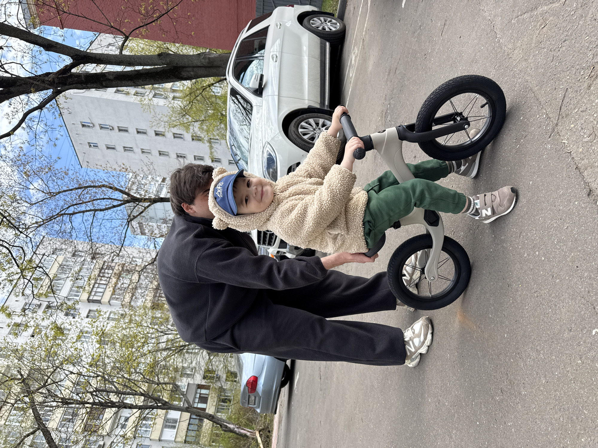 A young child wearing a beige sherpa-style hooded jacket and a blue baseball cap with a bear ear design is seated on a balance bike on an asphalt surface. The child is facing the viewer and holding the handlebars. Behind the child, a person in black clothing assists, holding the bike. A white SUV is parked behind the child, and a grey sedan is partially visible behind the adult. In the background, there is a multi-story building and trees with budding leaves. The scene is outdoors, and the location is Moscow, Russia.