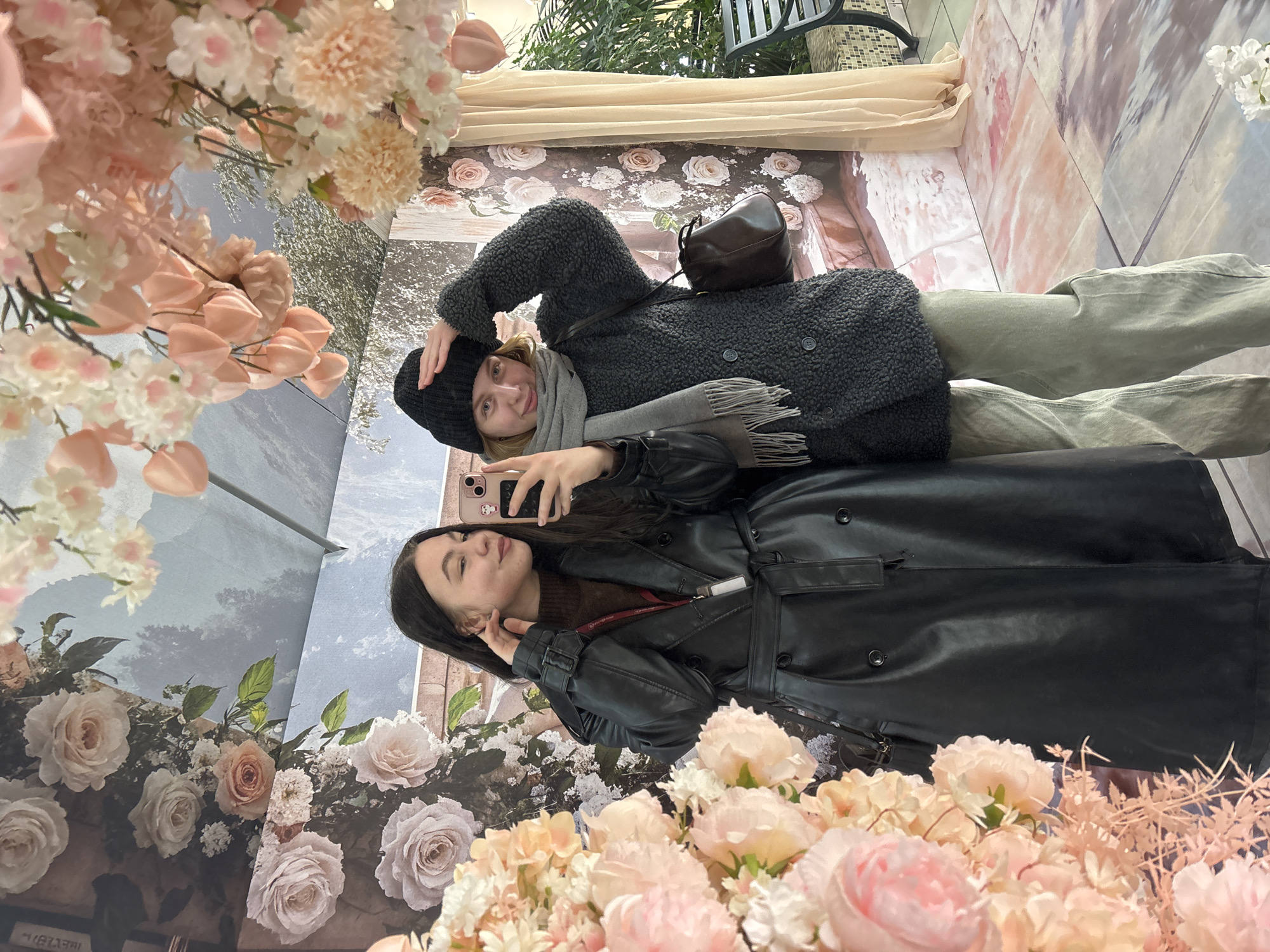 The image shows two young women taking selfies in an elaborately decorated indoor setting, likely a photo booth or an aesthetic public space. The scene is dominated by extensive artificial floral arrangements in shades of pink, peach, and white, creating a vibrant backdrop.
One woman, with blonde hair, is on the left side of the frame (if viewed upright). She wears a dark grey textured winter coat, a black beanie, a light grey scarf, and light green pants. A dark brown cross-body bag hangs over her shoulder. She smiles faintly, with one hand casually placed on her head, holding a phone with a pink case.
The second woman, with long dark hair, is on the right. She is dressed in a stylish black faux leather trench coat over a dark brown top. She holds her phone, also with a pink case featuring a character sticker, in a selfie pose, making a playful duck face and touching her cheek.
The background behind them consists of large decorative panels. One panel depicts a serene landscape with a cloudy sky and trees, while another features a pattern of light pink roses against a darker, possibly rustic, surface. Further floral clusters, including peonies and various small blossoms, are arranged both as foreground elements and hanging decorations, adding depth and a dreamlike quality to the setting. A draped, light-colored fabric also contributes to the decorative environment. The overall atmosphere suggests a modern public space or a specialized photo zone, common in venues like malls or event spaces, potentially in Perm, Russia. There is no discernible text visible in the image.
